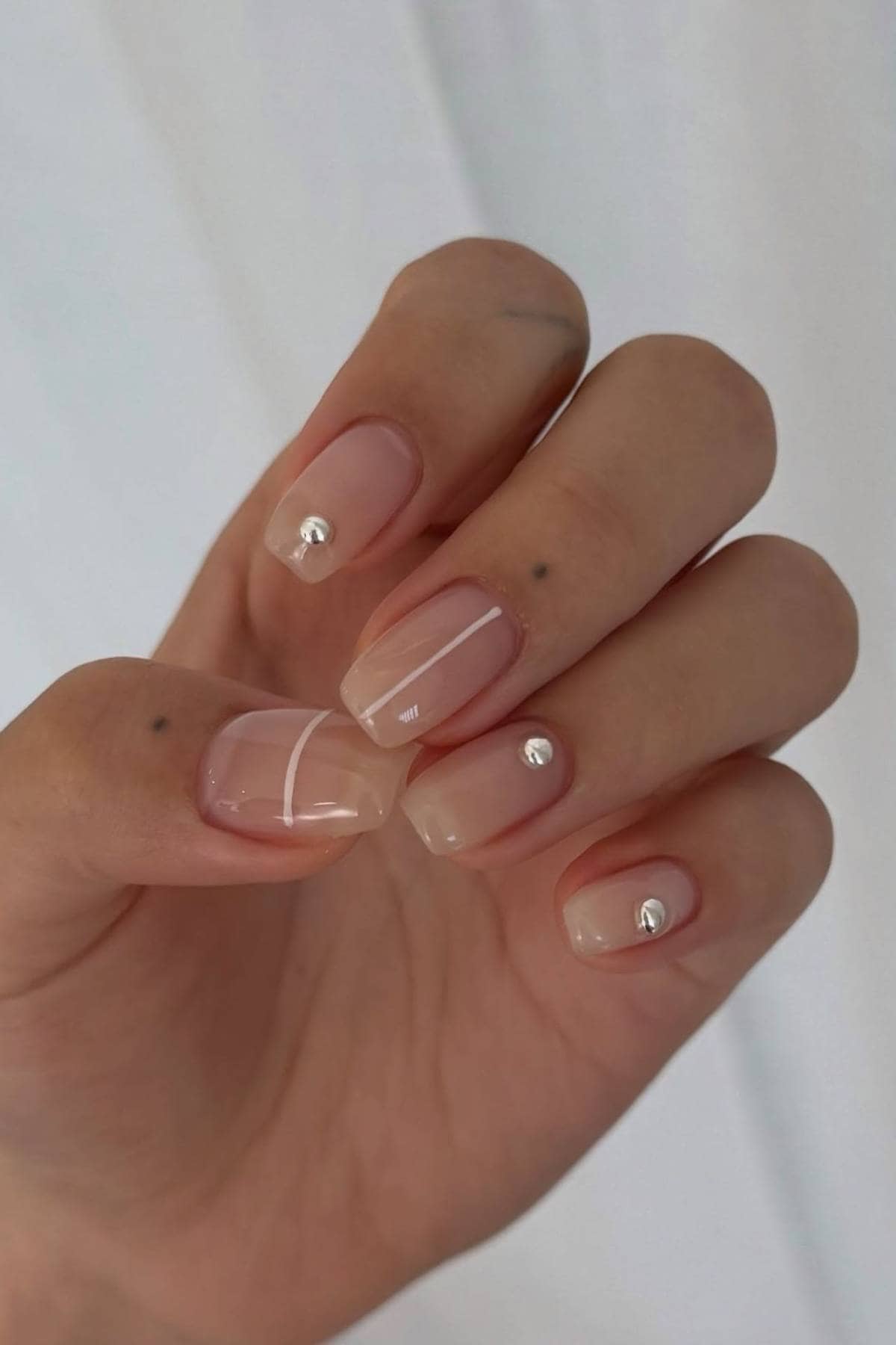 A hand with short, nude-colored, square-shaped nails featuring a glossy finish and small silver gems on each nail.