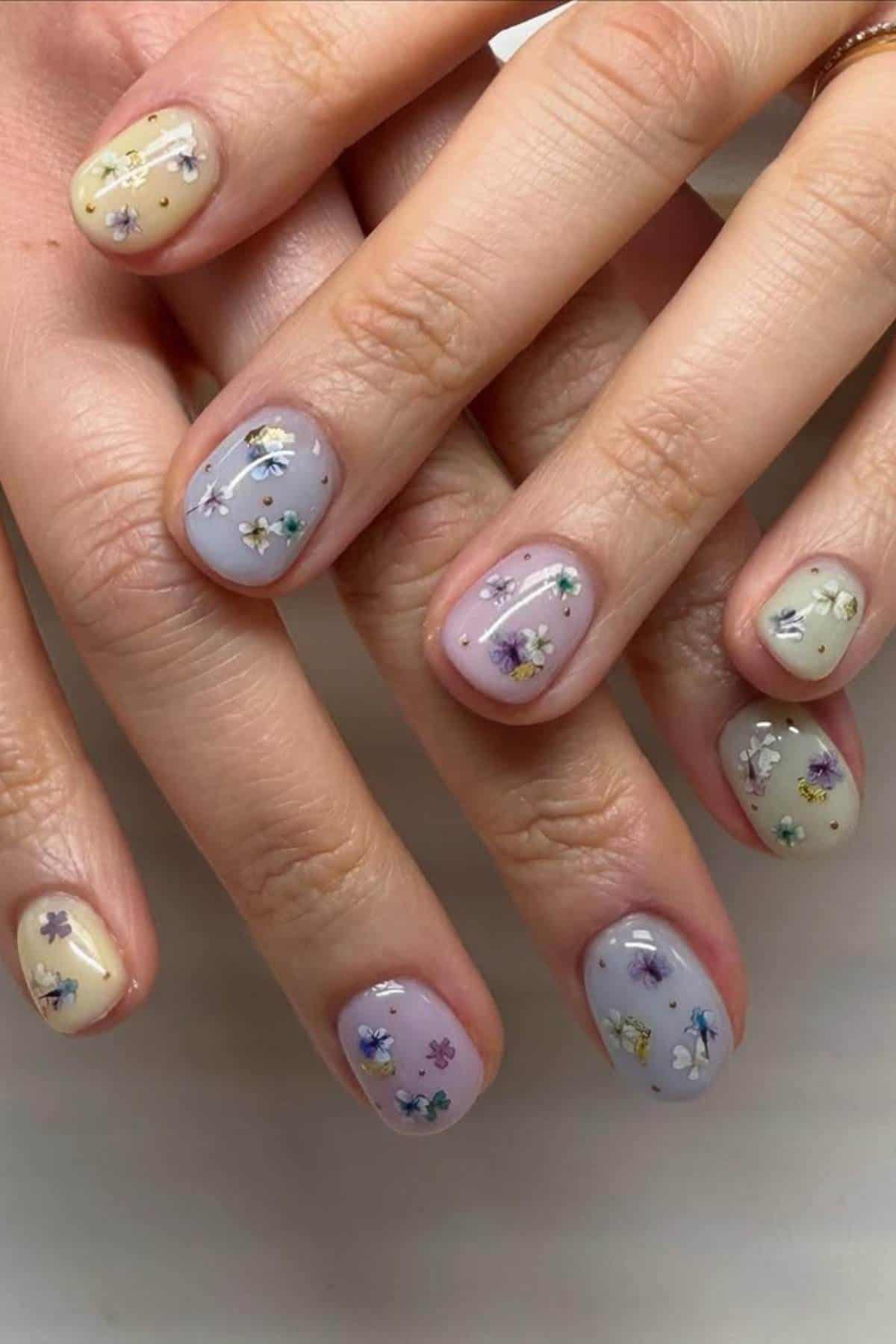 Close-up of hands with short nails painted in pastel yellow and lavender, decorated with small floral designs on each nail.