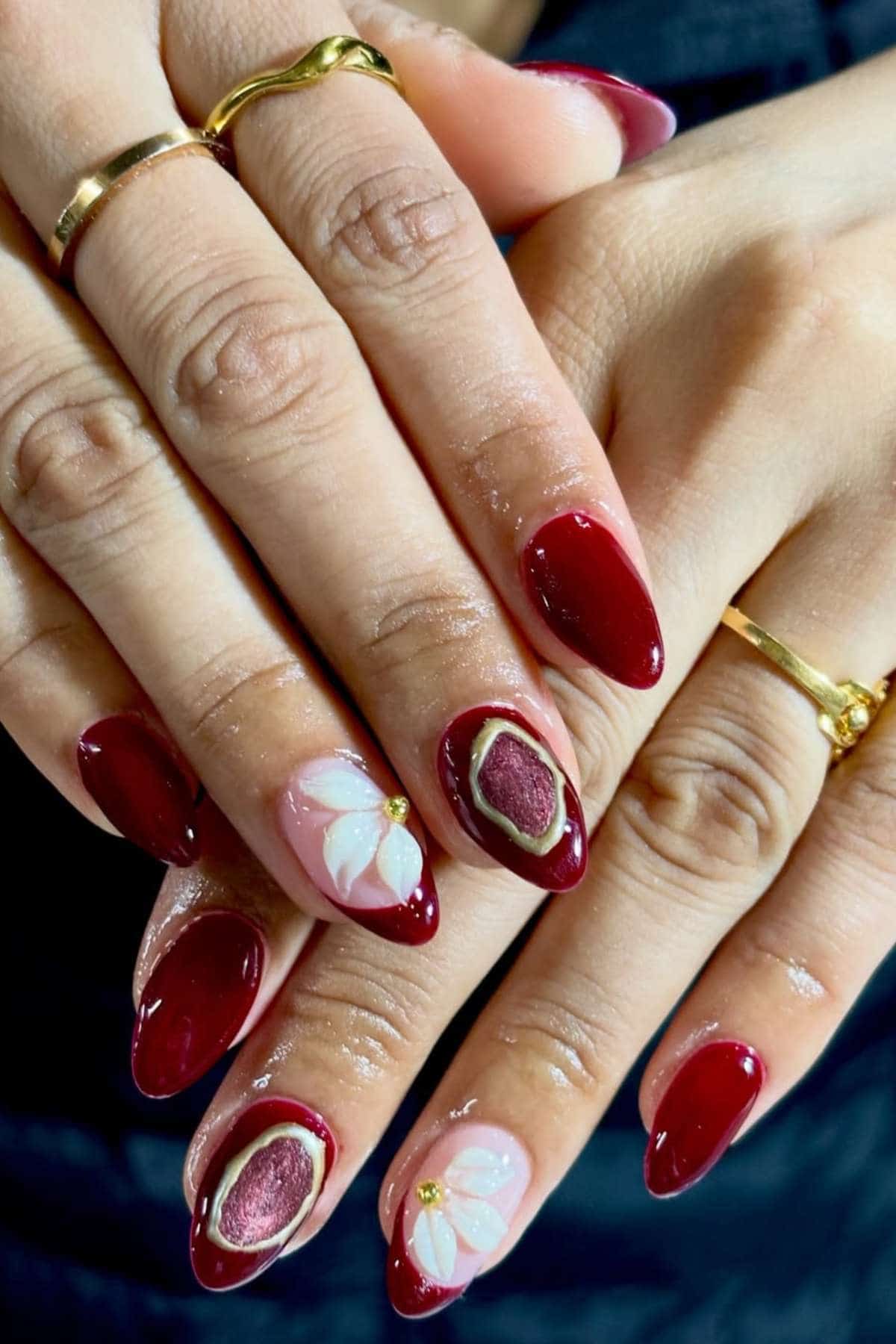 Hands with almond-shaped nails painted dark red, featuring two accent nails with white flower designs and gold ring details. Gold rings are worn on two fingers.