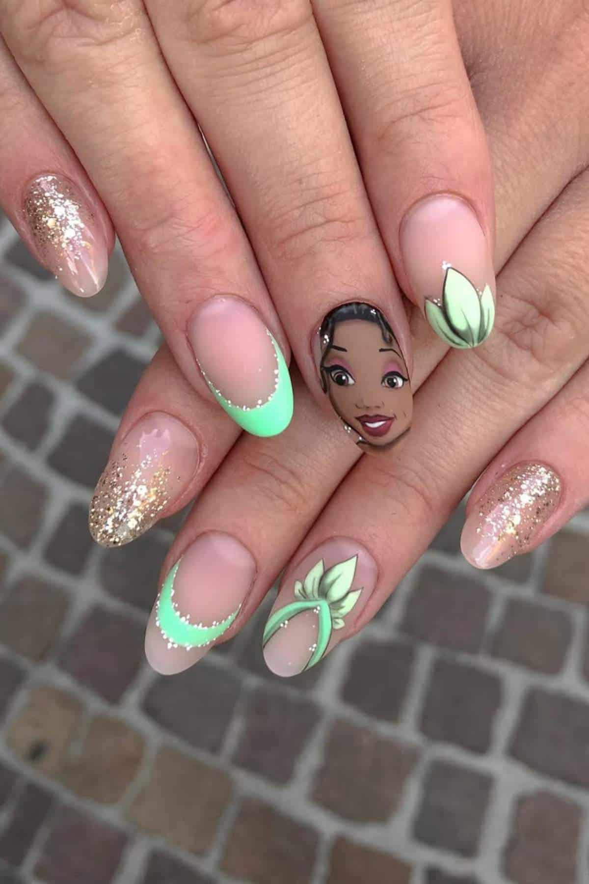 Close-up of hands with manicured nails featuring green tips, gold glitter, lotus flower designs, and one nail with a painted face of a woman.