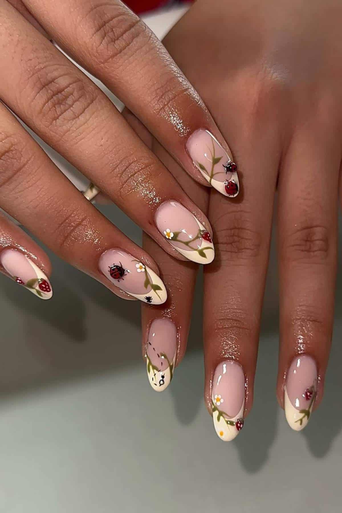 Hands with almond-shaped nails featuring a nude base, floral designs, and small ladybug nail art accents on each nail.