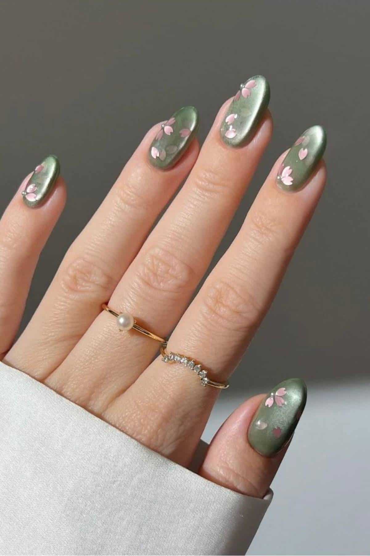 A hand with green, almond-shaped nails featuring small pink flower designs, wearing two gold rings, one with a pearl and one with small gems.