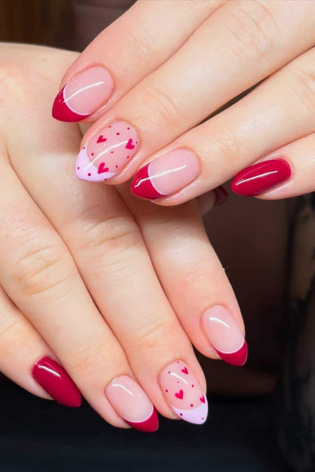 Hands with almond-shaped nails featuring a pink and red French tip design; accent nails have red and pink heart patterns on a light pink base.
