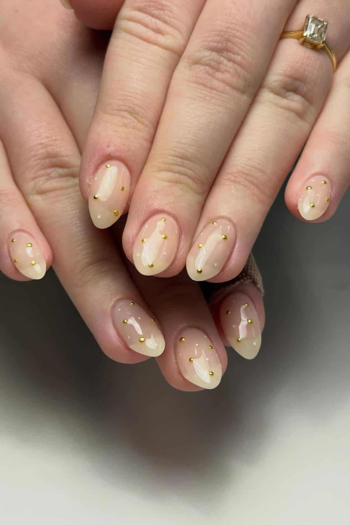 Hands with almond-shaped nails feature a nude gradient base and small gold dot accents. One hand has a gold ring with a rectangular stone on the ring finger.