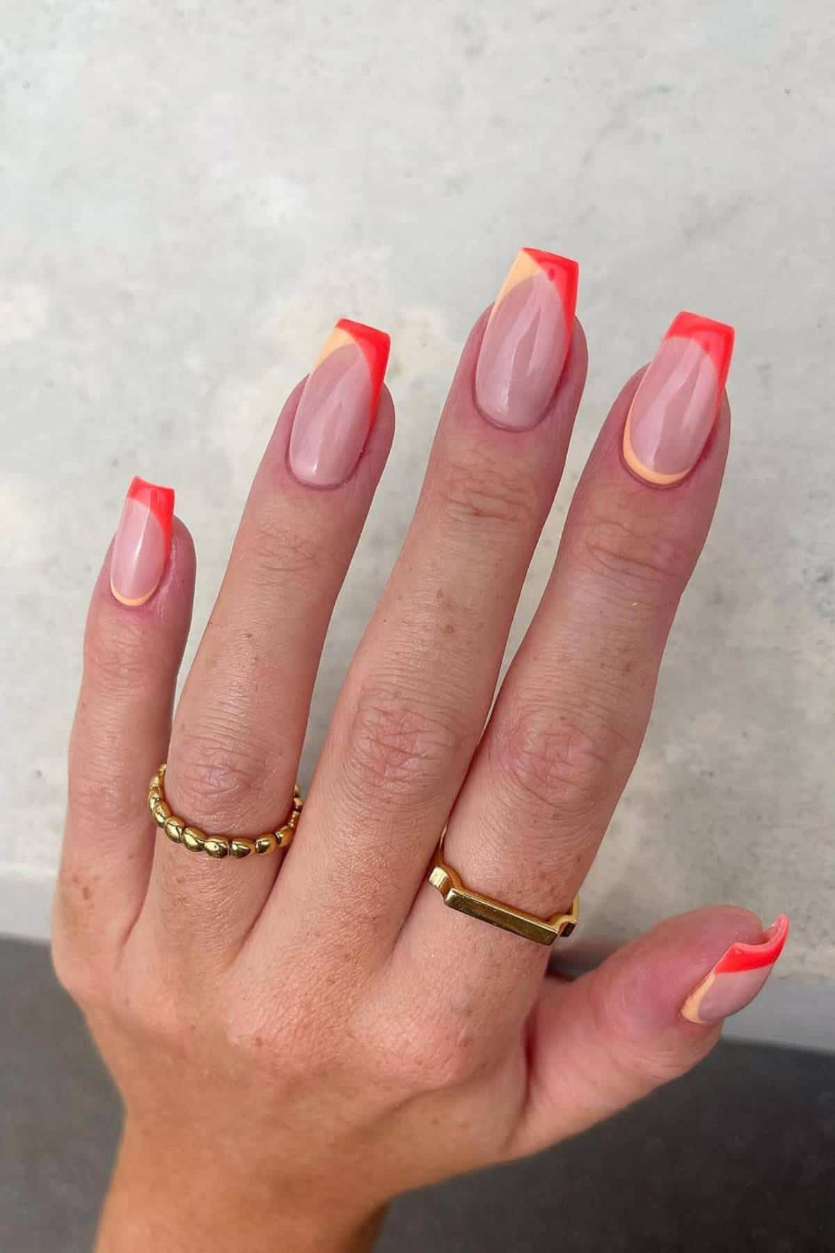 A hand with long, almond-shaped nails featuring a neon orange and yellow French tip design, wearing two gold rings.