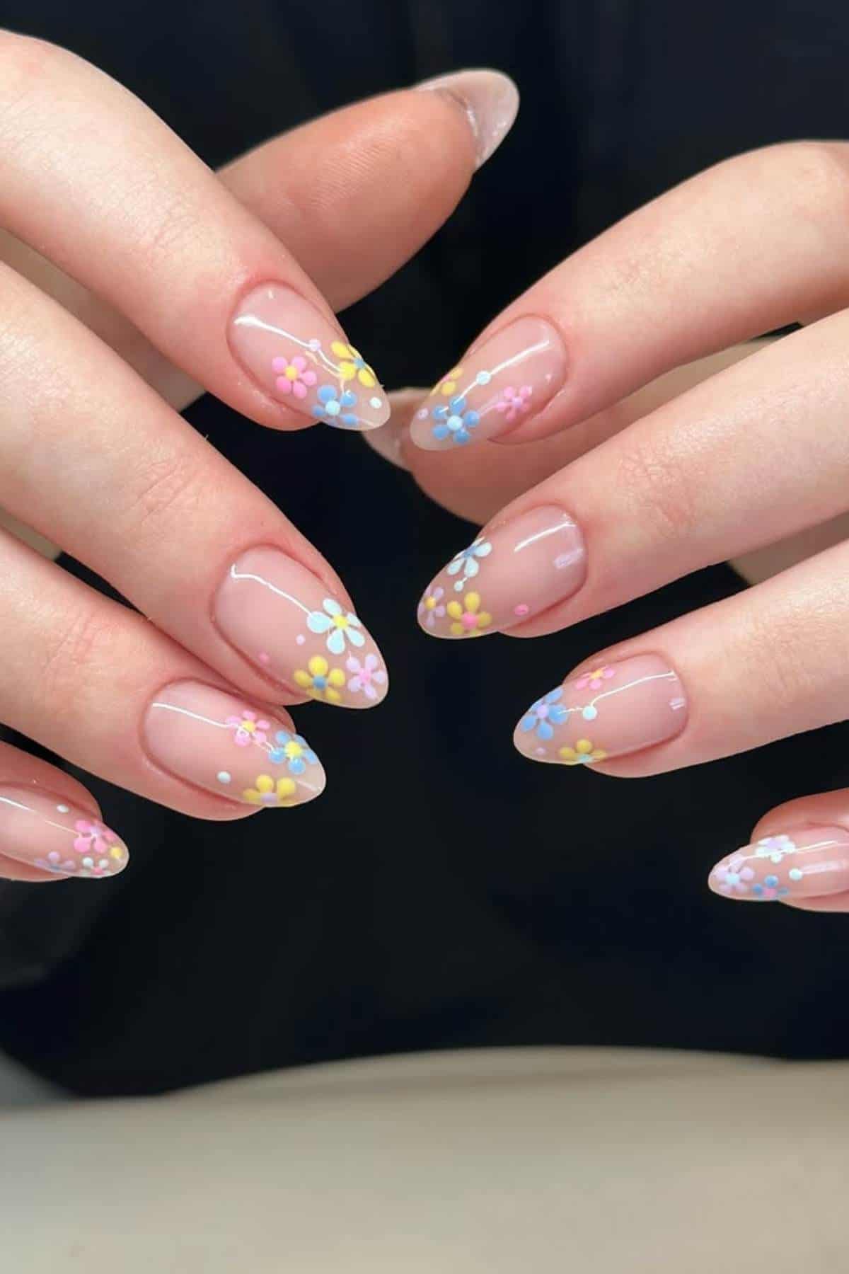 Close-up of hands with almond-shaped nails featuring a clear base and pastel-colored floral nail art in yellow, blue, and pink.
