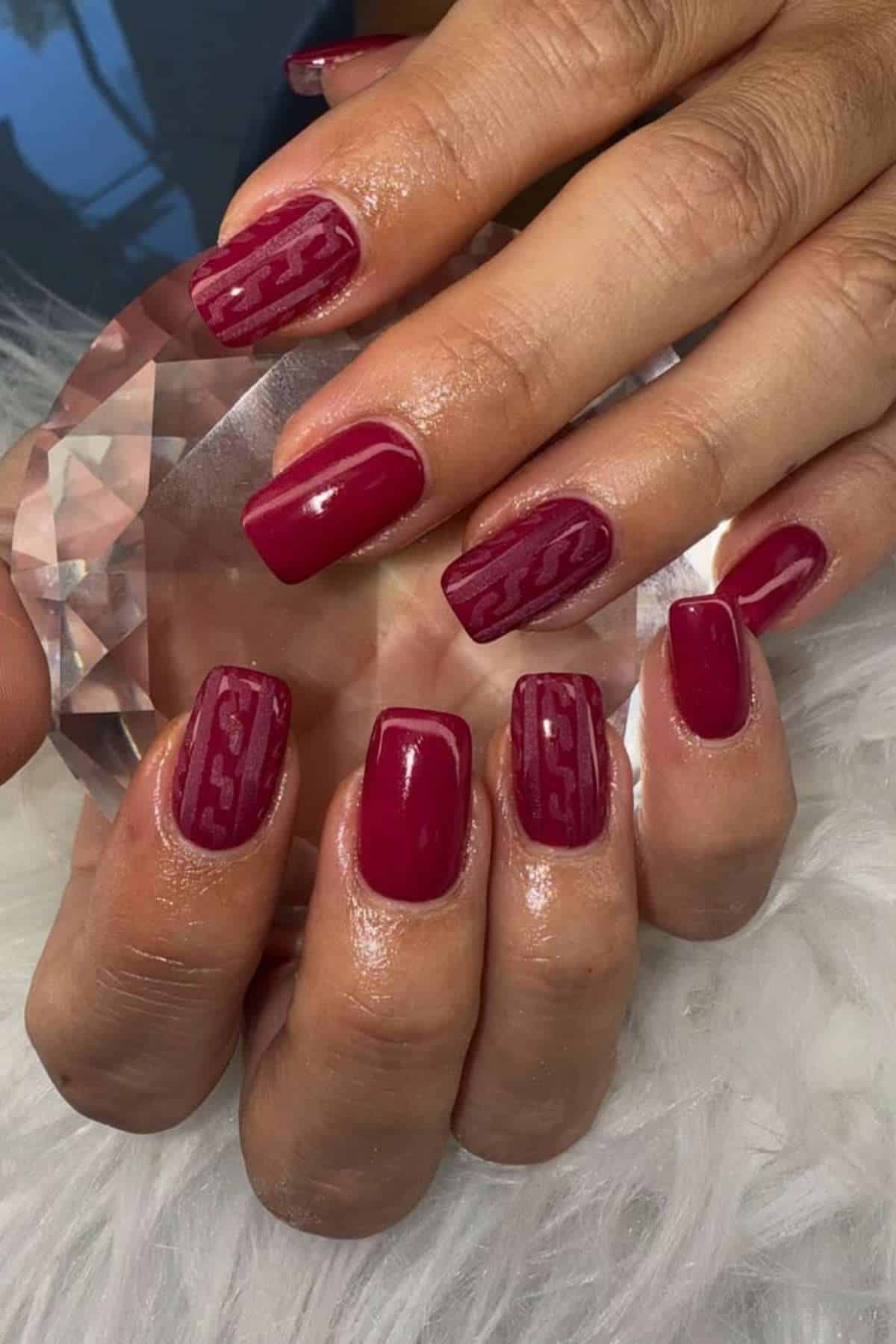 A hand with glossy, dark red square-shaped nails; some nails have a subtle wavy line pattern. The hand is holding a faceted clear object, resting on a white fuzzy surface.