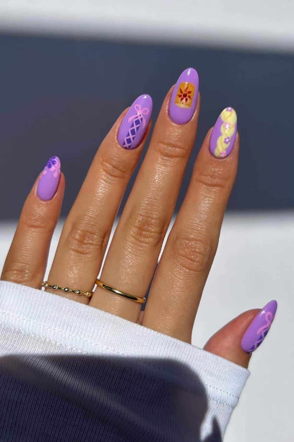 A hand with almond-shaped nails painted in purple tones, featuring varied designs including florals, hearts, and geometric patterns on each nail.