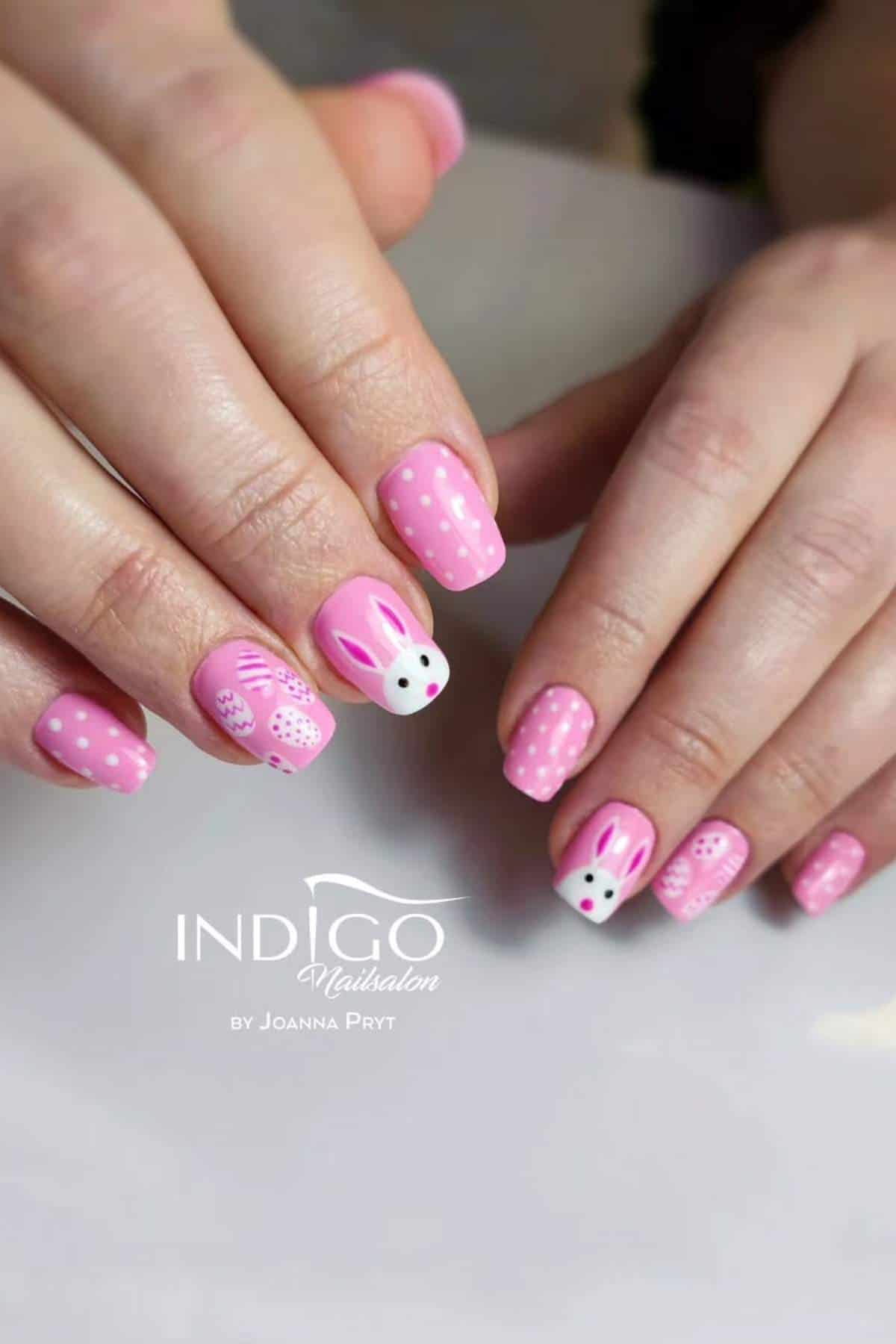 Two hands with pink nail polish featuring white polka dots and bunny face designs on select nails. The background is neutral and the Indigo Nailsalon logo is visible.