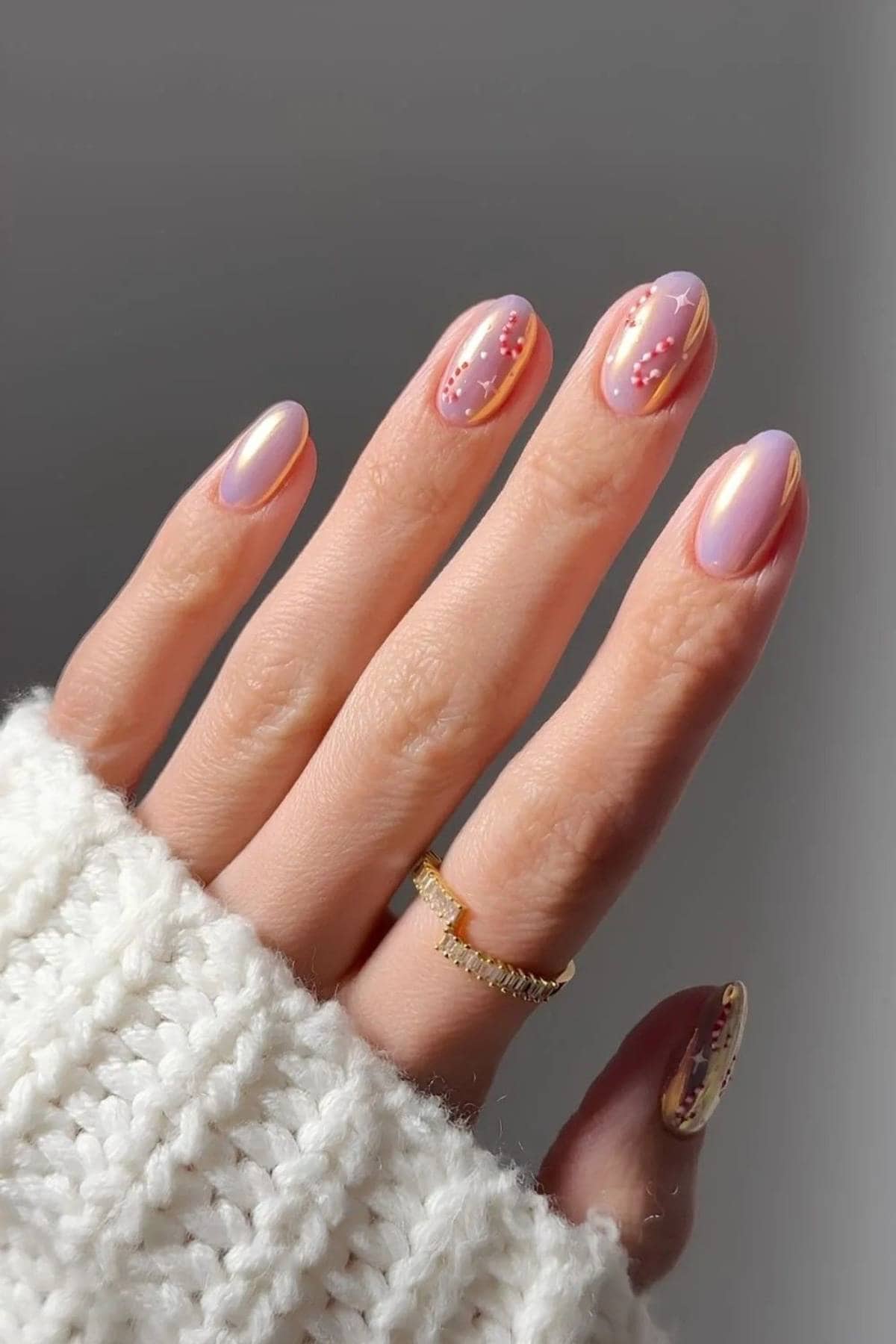 A hand with light pink, glossy manicured nails featuring small dot designs and gold accents, wearing a gold ring and a white knit sweater.