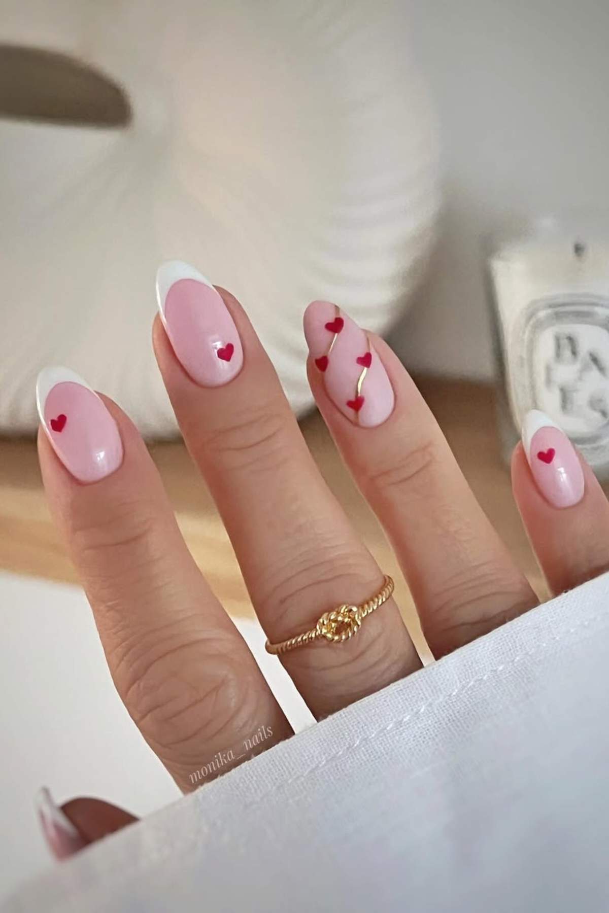A hand with long, almond-shaped nails painted pink, featuring white tips and small red heart designs, is shown with a gold ring on the ring finger.
