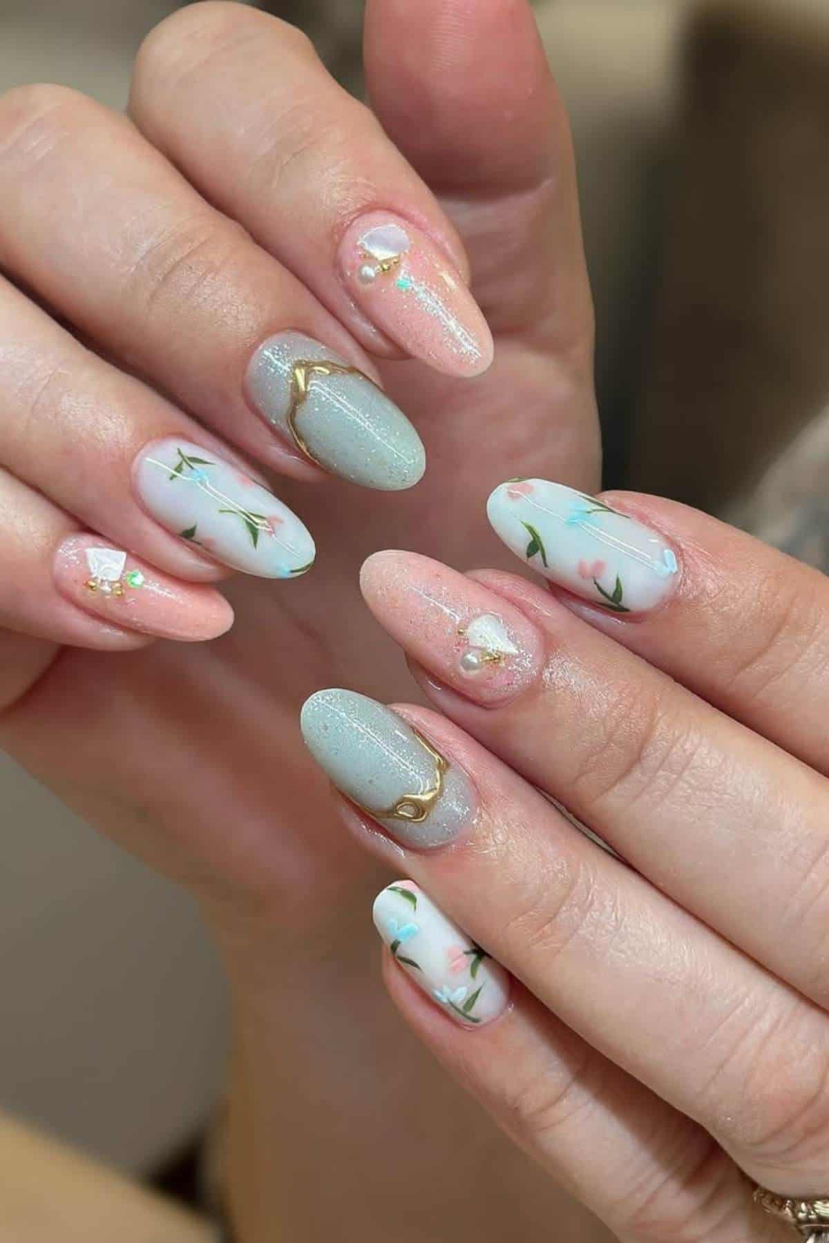 Hands with almond-shaped nails featuring pastel blue and pink polish, floral designs, gold accents, and small embellishments.