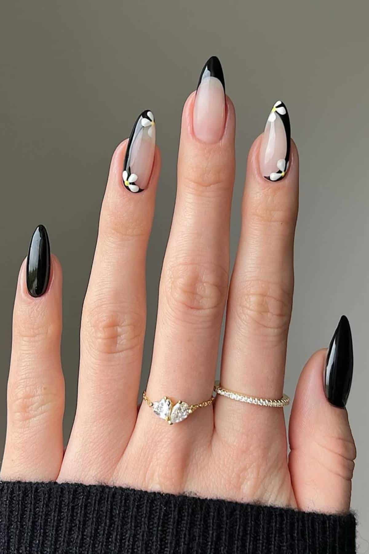 A hand with almond-shaped nails painted black, with three nails featuring minimalist white daisy designs near the cuticle; person wears two delicate rings.
