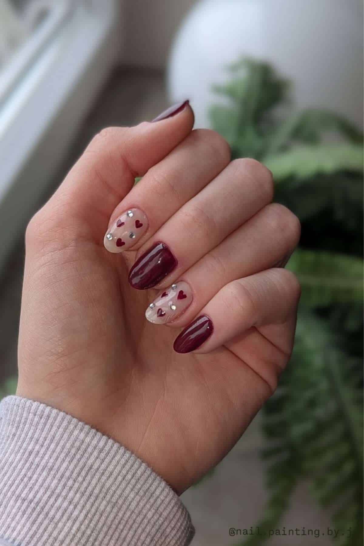 A hand with manicured nails; some nails are painted burgundy and others are clear with small red heart designs and silver dots. A plant and window are in the background.