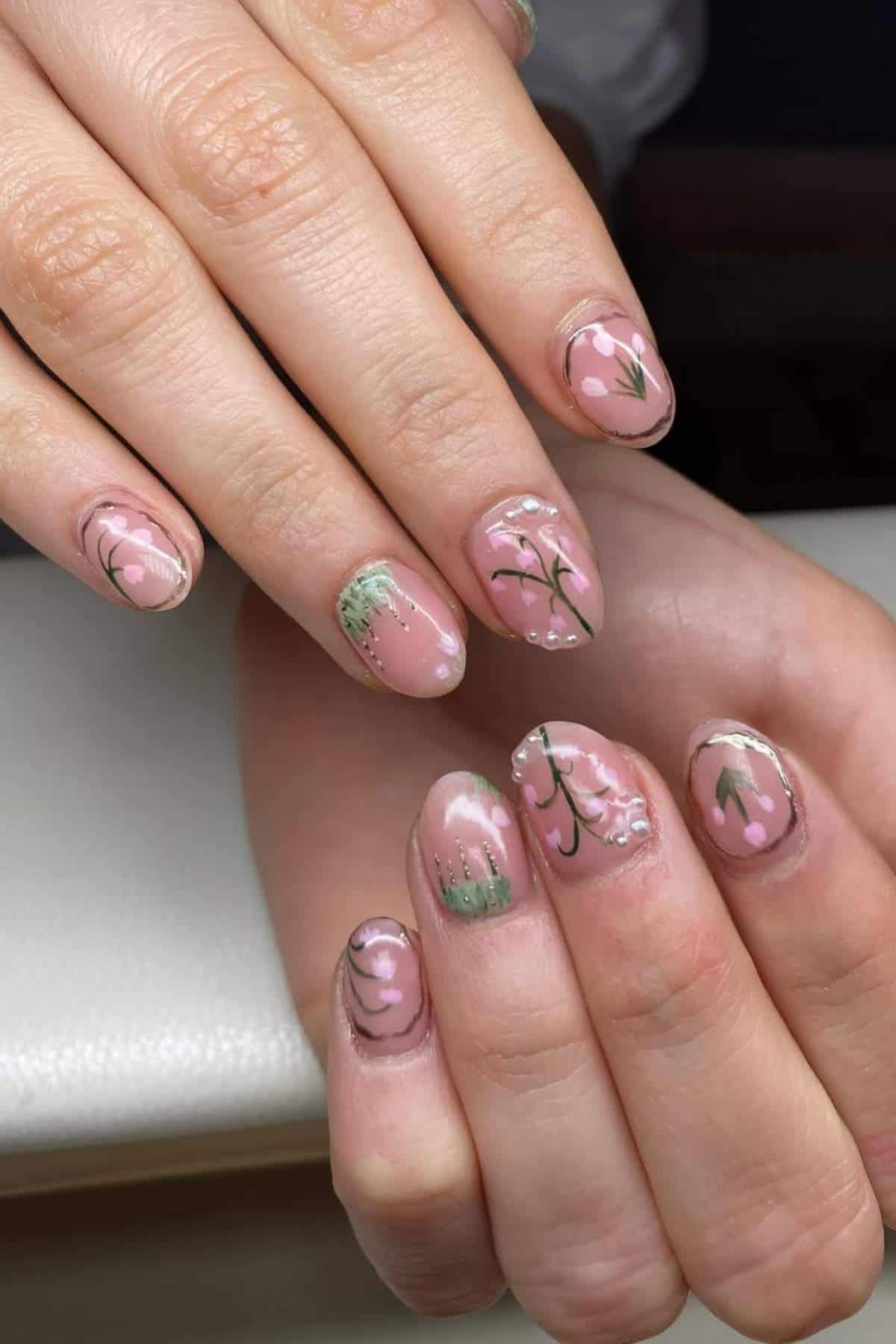 Hands with short, almond-shaped nails painted with light pink polish and decorated with green leaves and white floral designs.