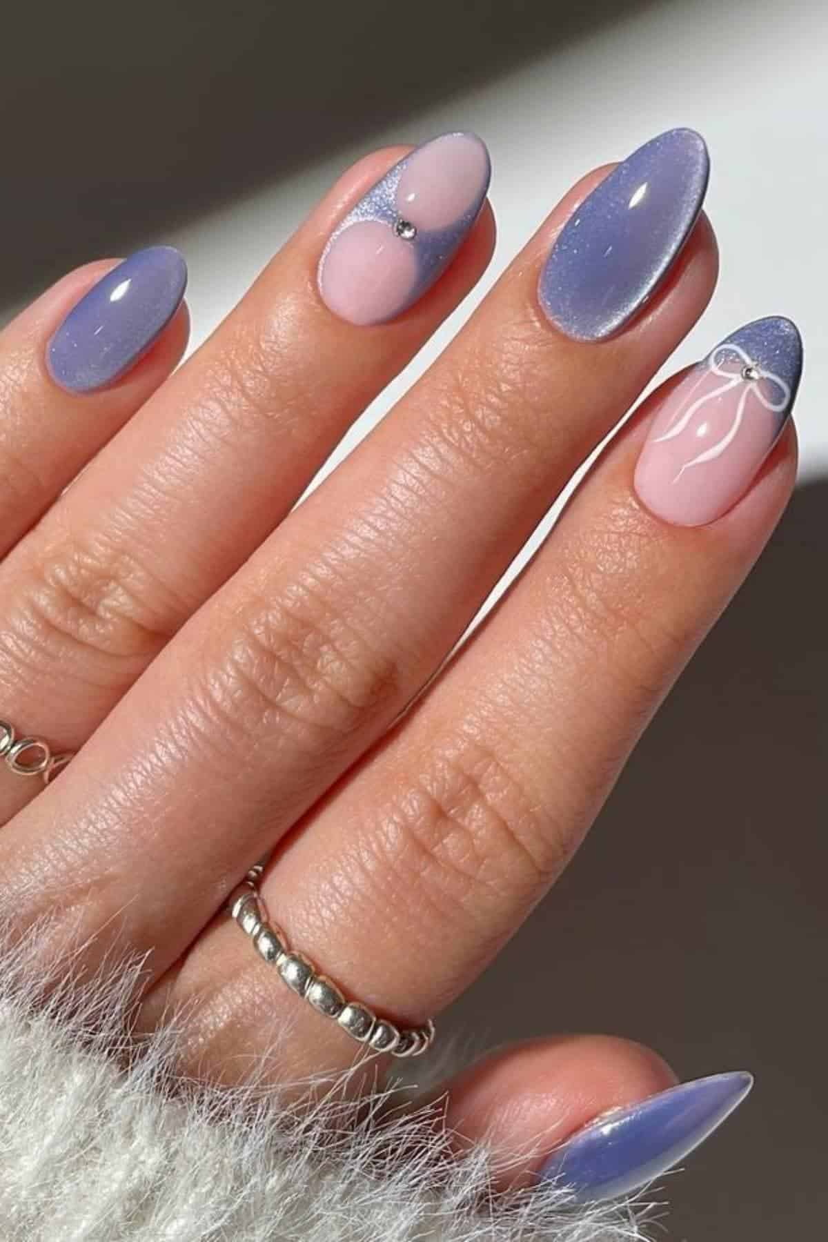 A hand with almond-shaped nails featuring a pastel purple and pink manicure, some nails accented with tiny rhinestones and white line art, and silver rings on two fingers.