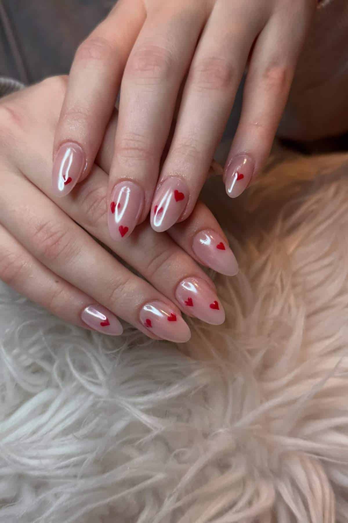 Hands with almond-shaped nails painted in a nude color, each nail decorated with small red heart designs, resting on a white, fluffy surface.