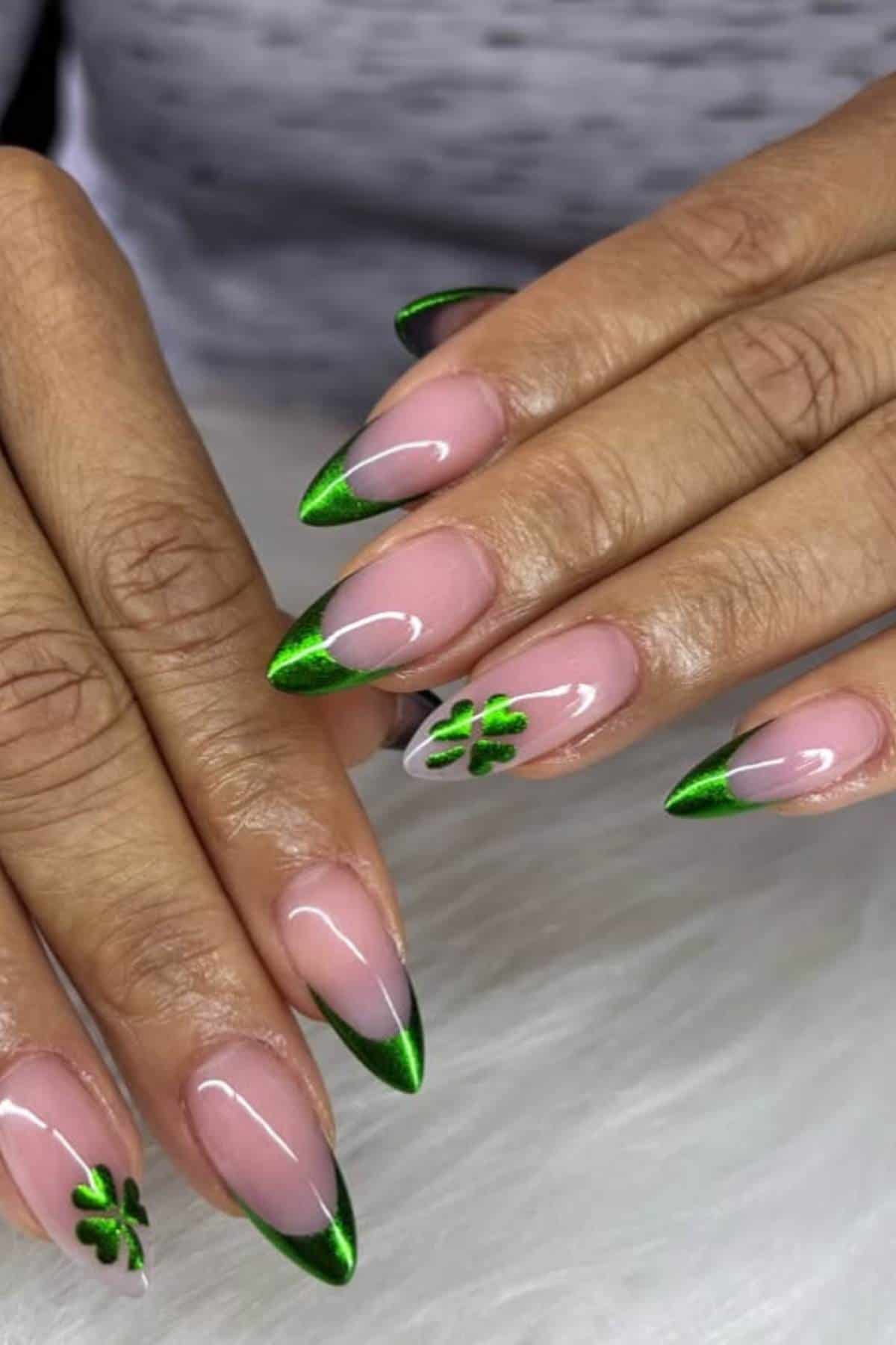 Hands with almond-shaped nails featuring green metallic French tips and two accent nails with painted green four-leaf clovers on a light background.