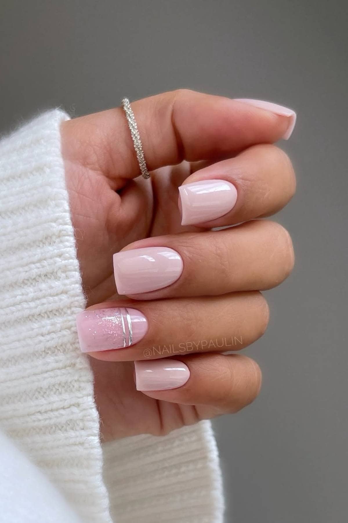 A hand with short, square-shaped nails painted light pink, with the ring finger featuring glitter and two thin silver lines. The person wears a silver ring and a white sweater.