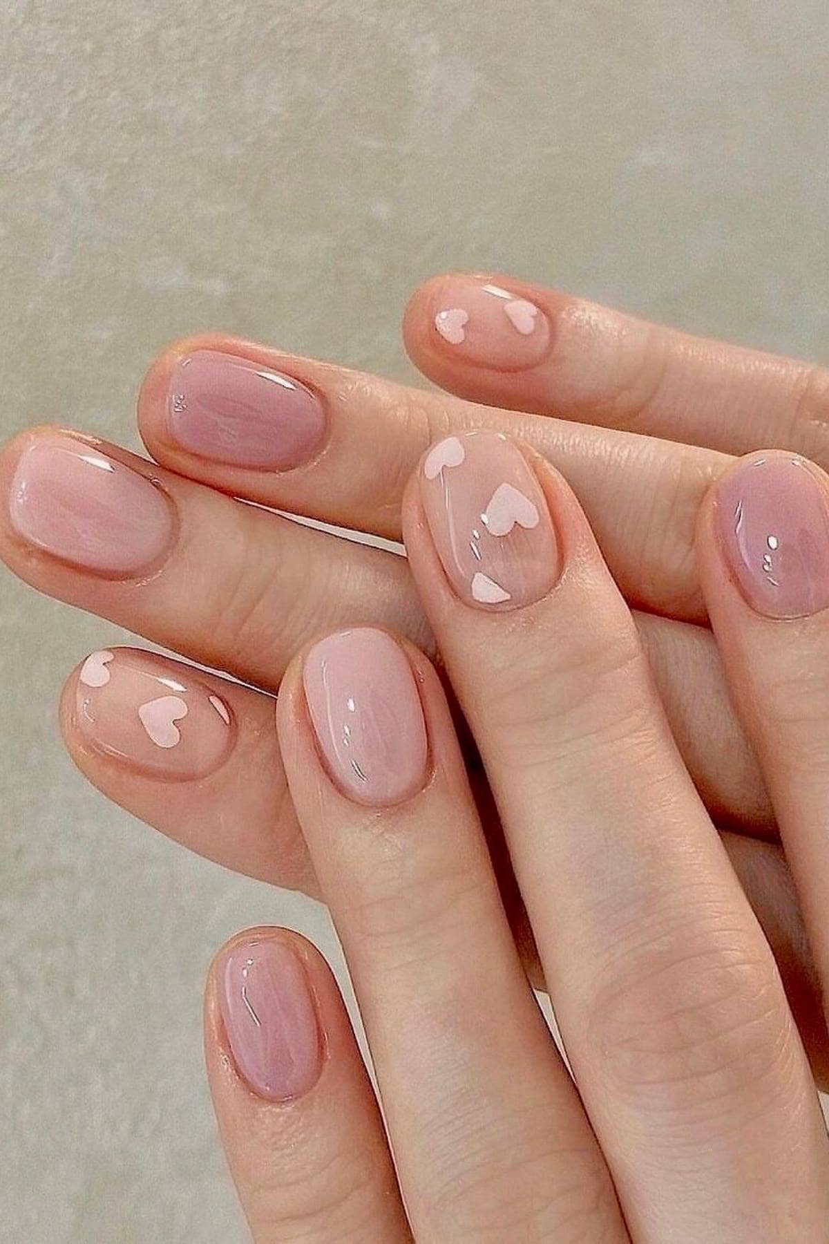 Close-up of hands with short, oval-shaped nails painted in light pink polish; some nails feature small white heart designs.