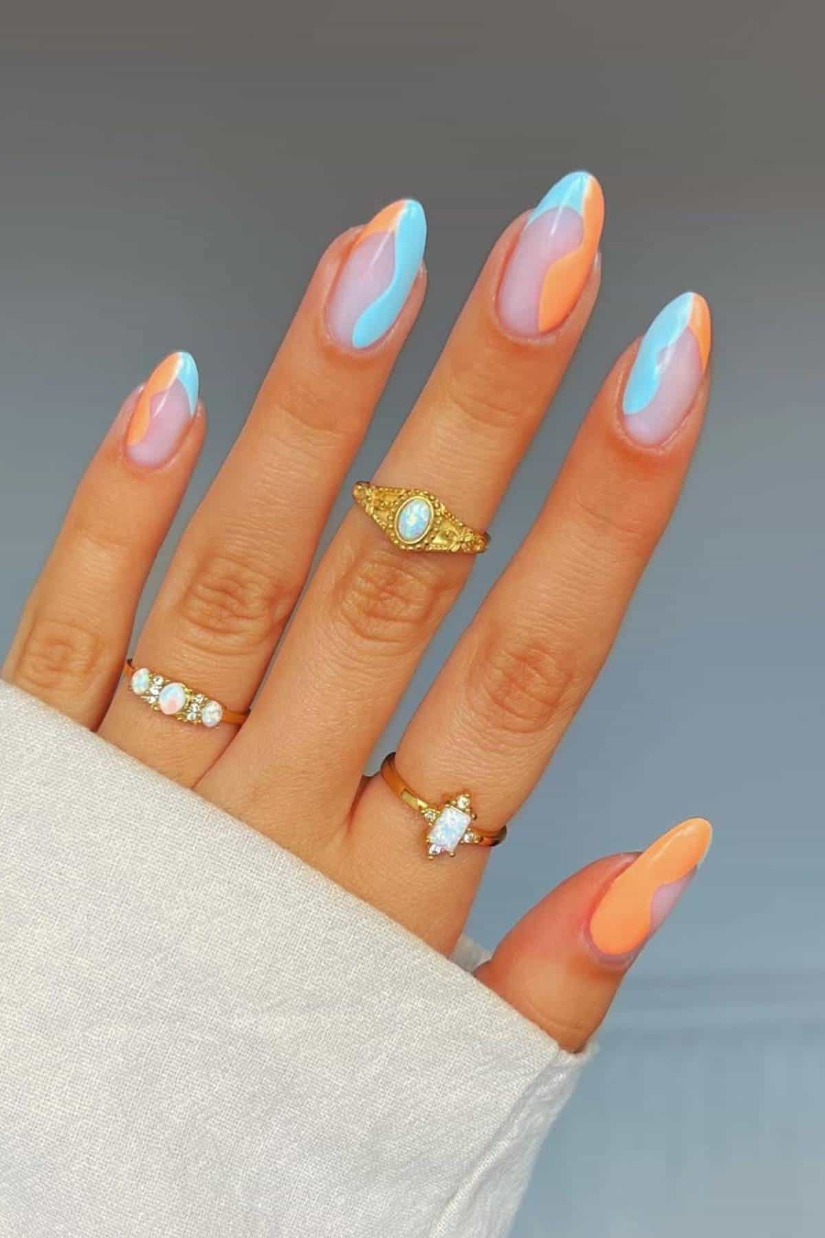 A hand with long, almond-shaped nails features a pastel blue, pink, and orange abstract design. The fingers wear three gold rings with gemstones.