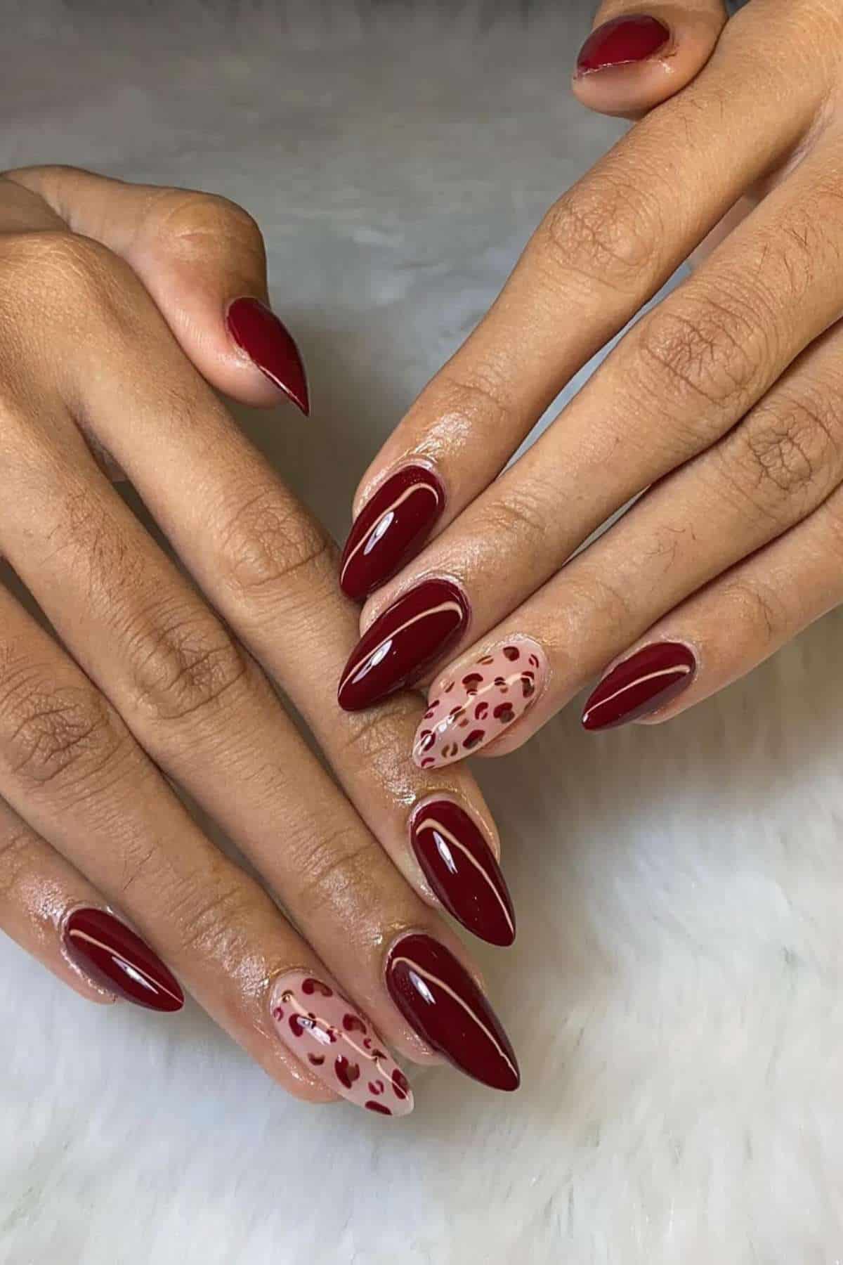 Hands with almond-shaped nails painted dark red, featuring leopard print designs on the ring fingers over a nude base, resting on a white textured surface.