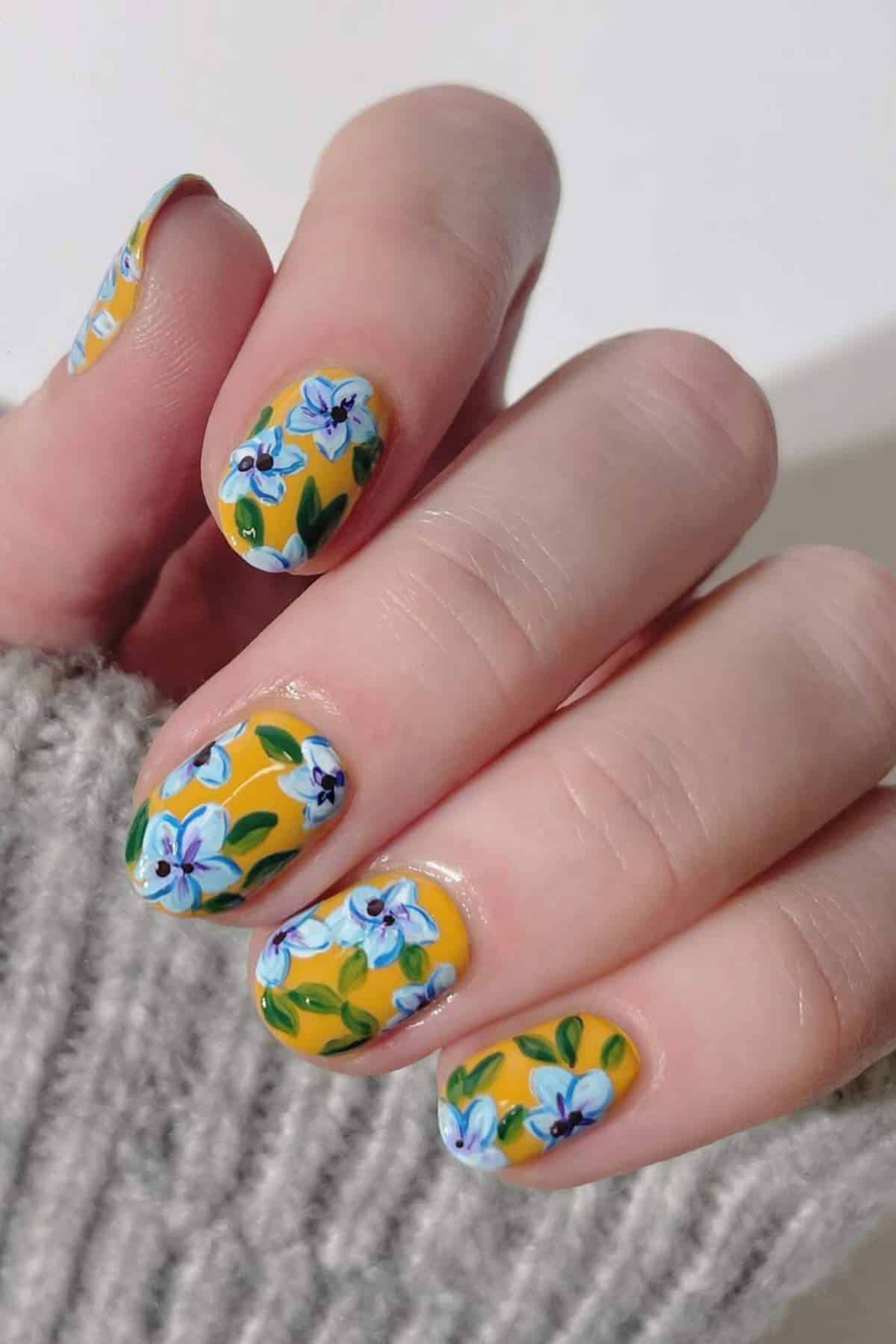 A hand with short nails painted yellow and decorated with detailed blue, white, and green floral designs. The person is wearing a light gray knit sweater.