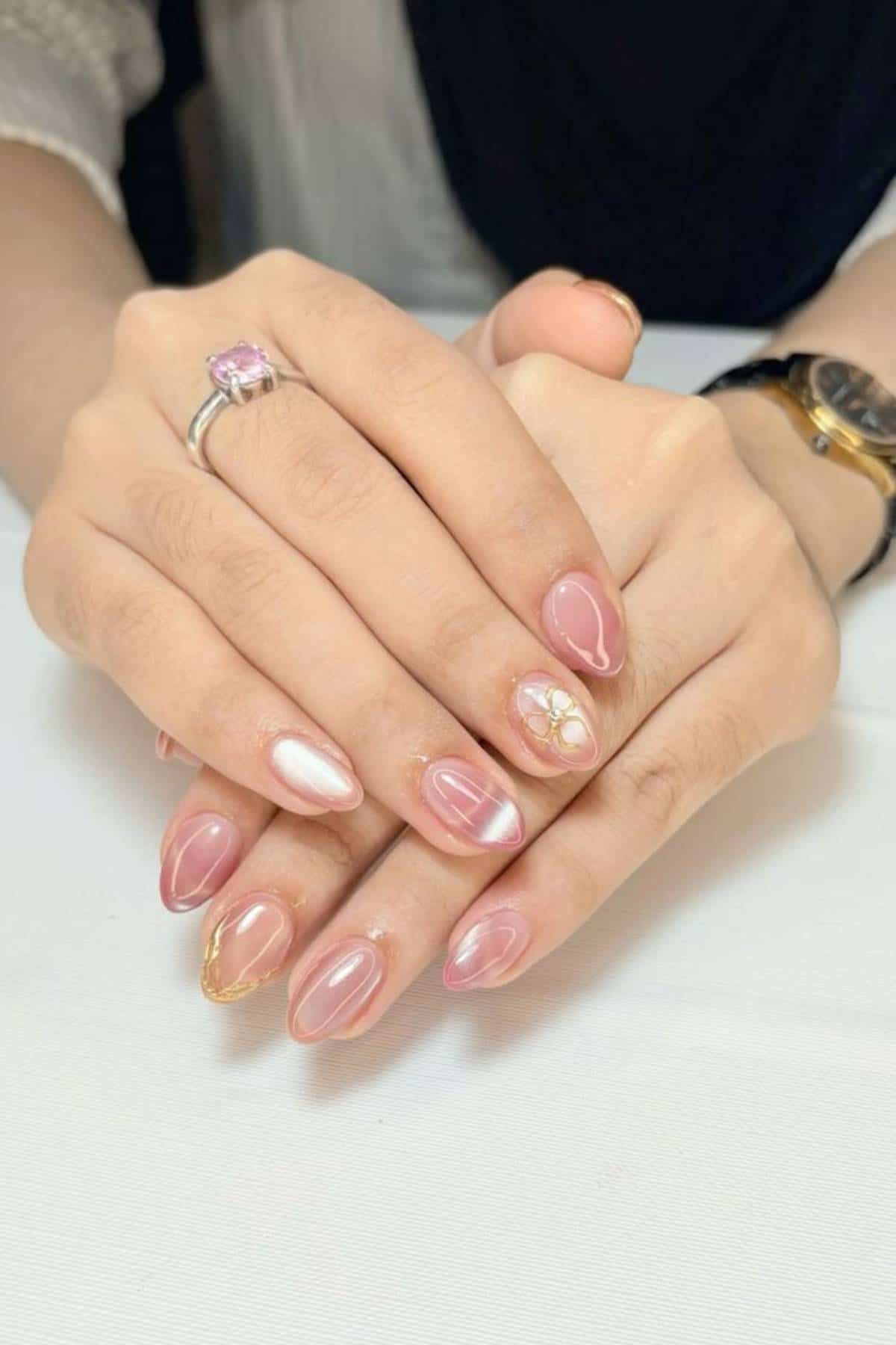 Hands with manicured nails featuring pink and white designs, gold accents, and floral patterns; a pink gemstone ring and a watch are visible.