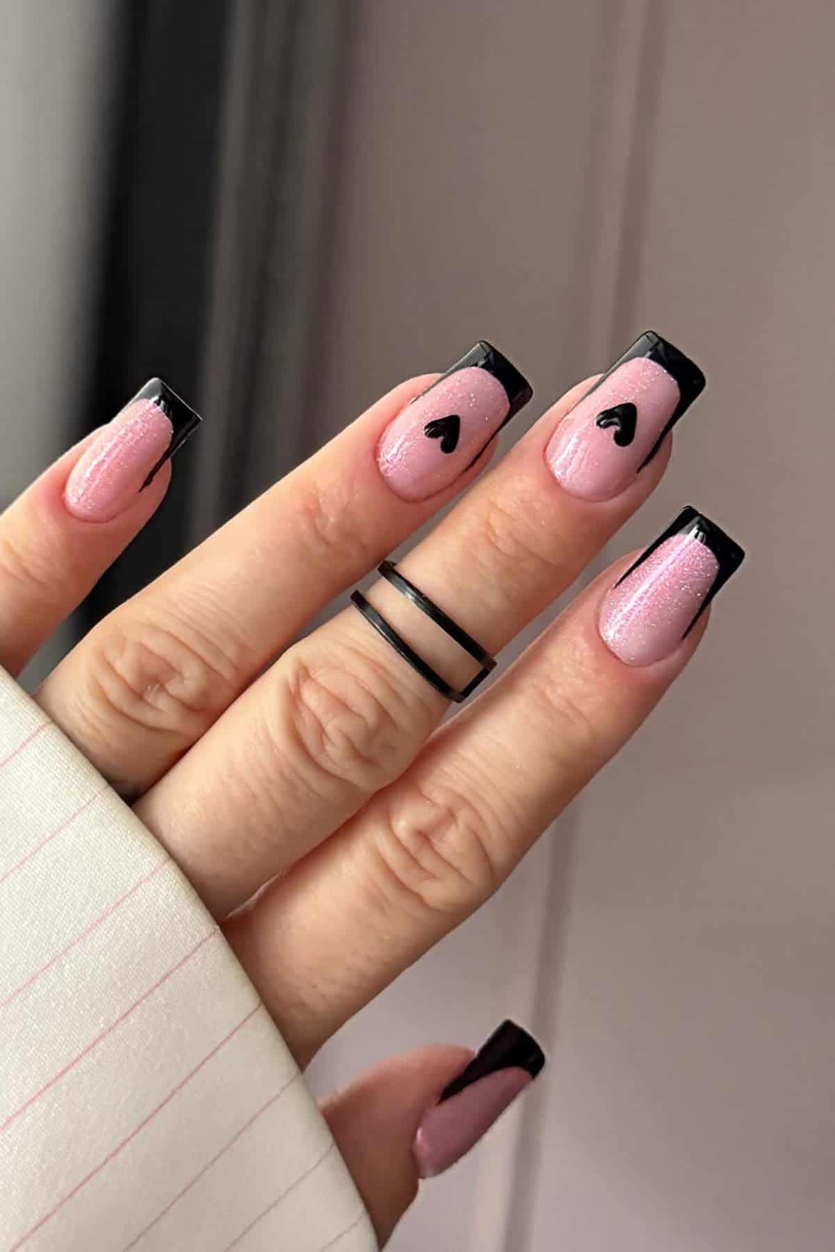 A hand with pink nails featuring black French tips and small black heart designs on the index and middle fingers. The person is wearing a double band black ring.