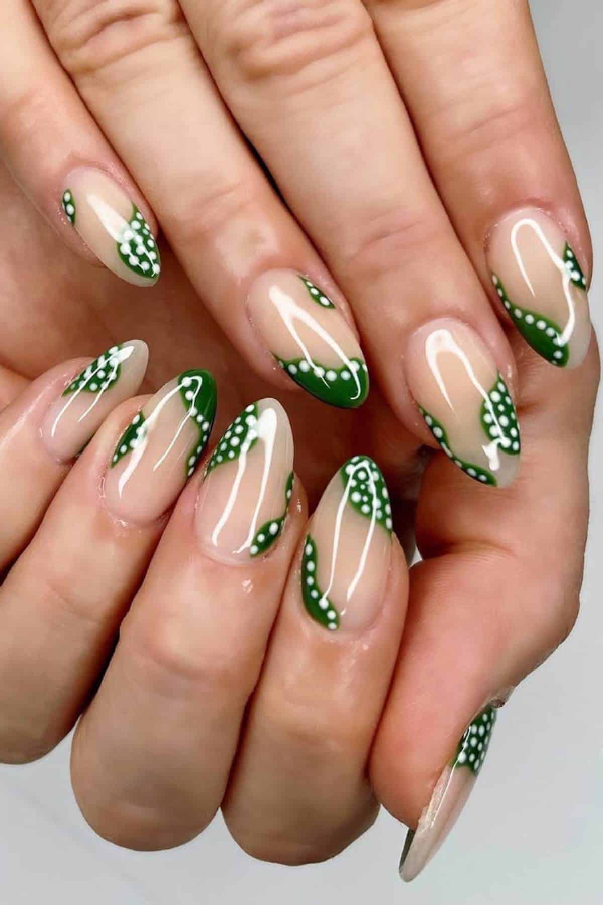 Hands with almond-shaped nails featuring a glossy nude base, green tips, and small white dot accents.