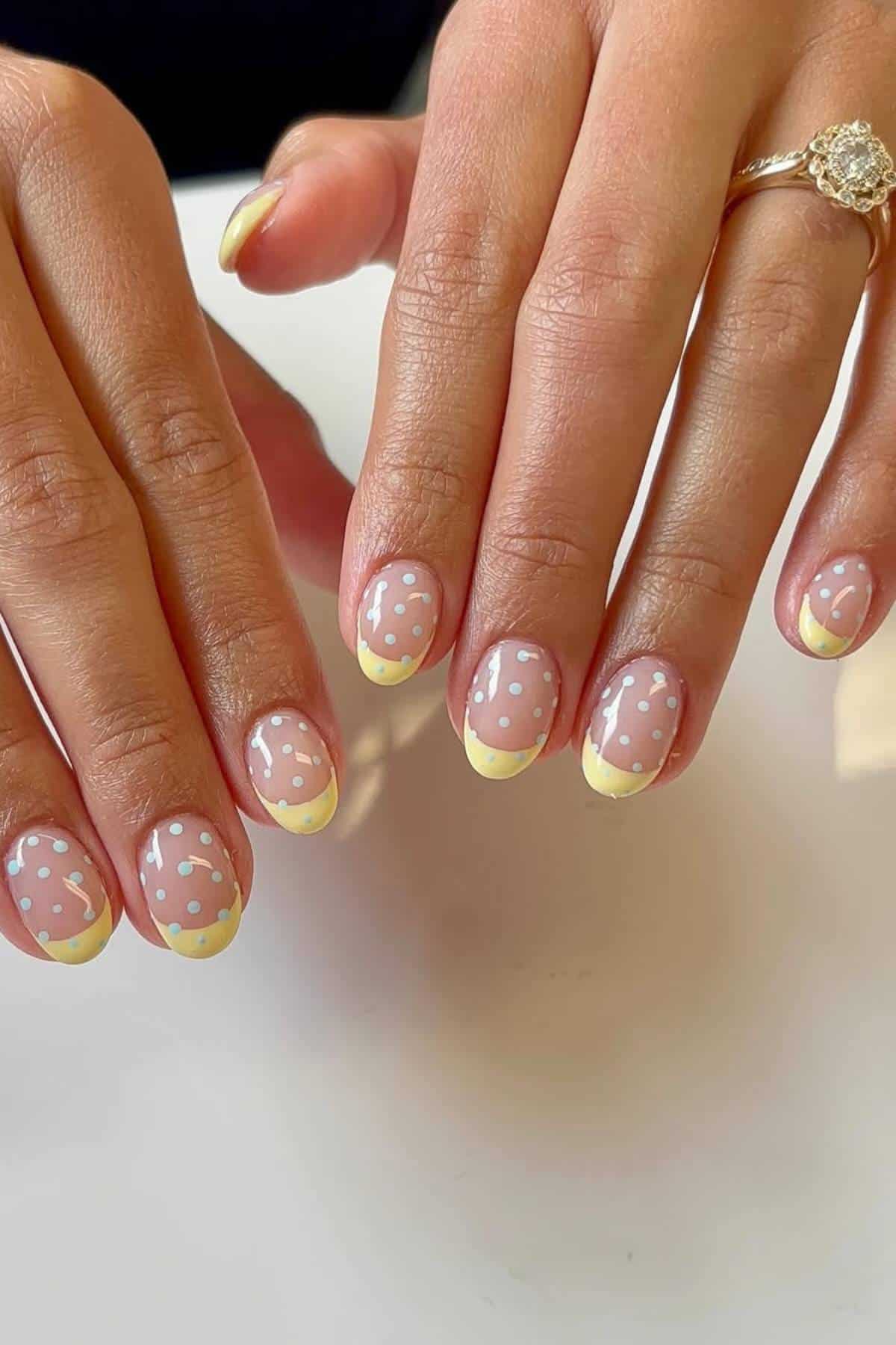 Hands with short nails featuring yellow French tips and white polka dot nail art, shown against a white background. One hand has a gold ring with a round stone.
