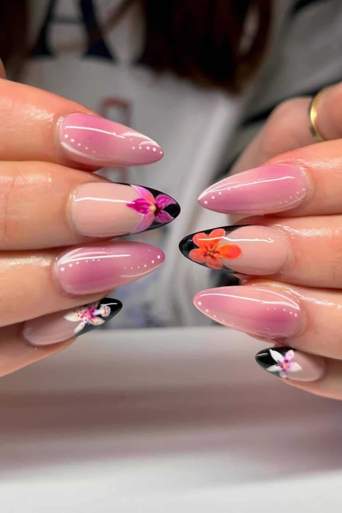 Close-up of hands with almond-shaped nails featuring a pink and nude base, black French tips, and detailed floral designs in pink, red, and purple tones.