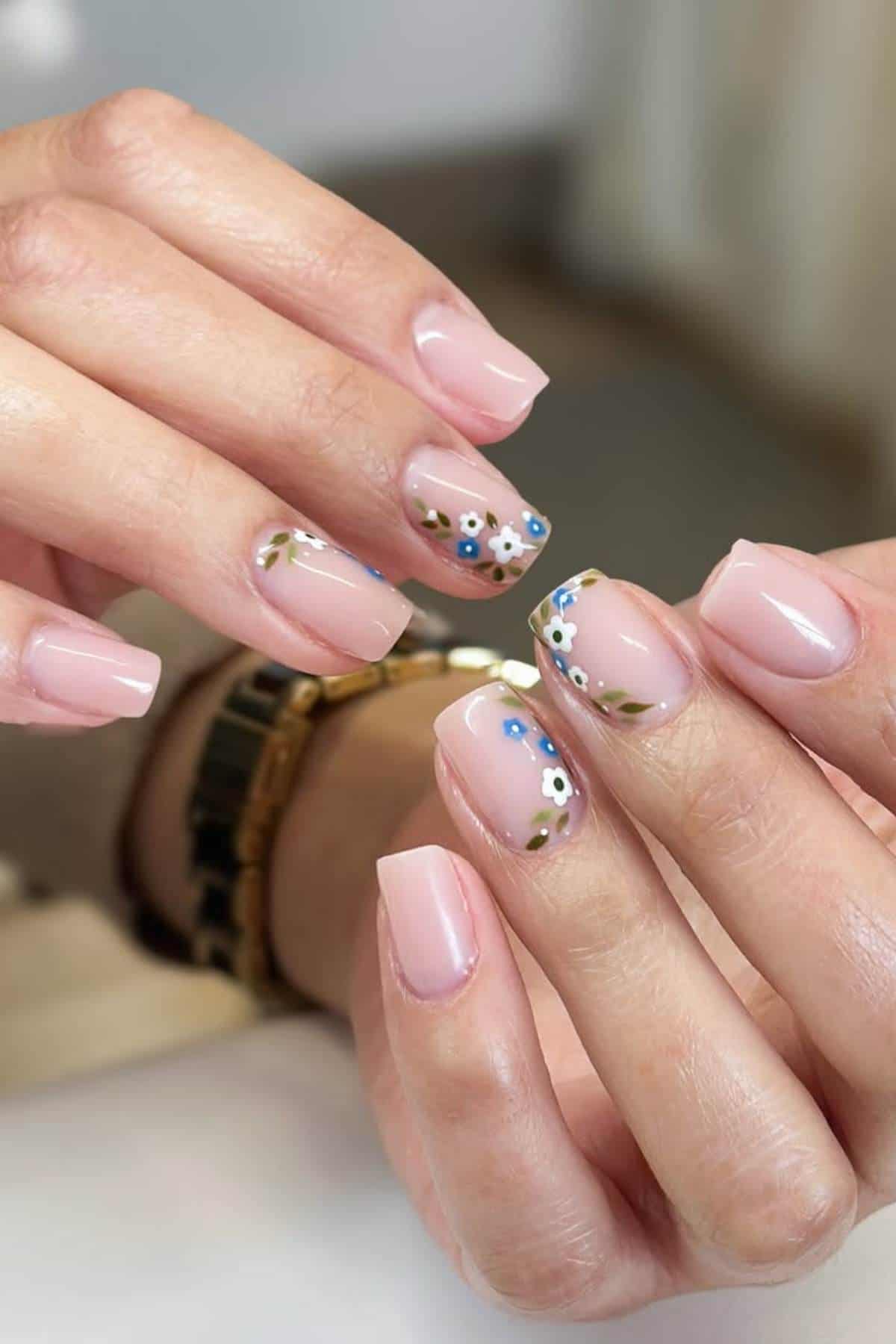 Hands with square-shaped nails painted in a light pink color; some nails feature floral nail art with small blue and white flowers and green leaves.