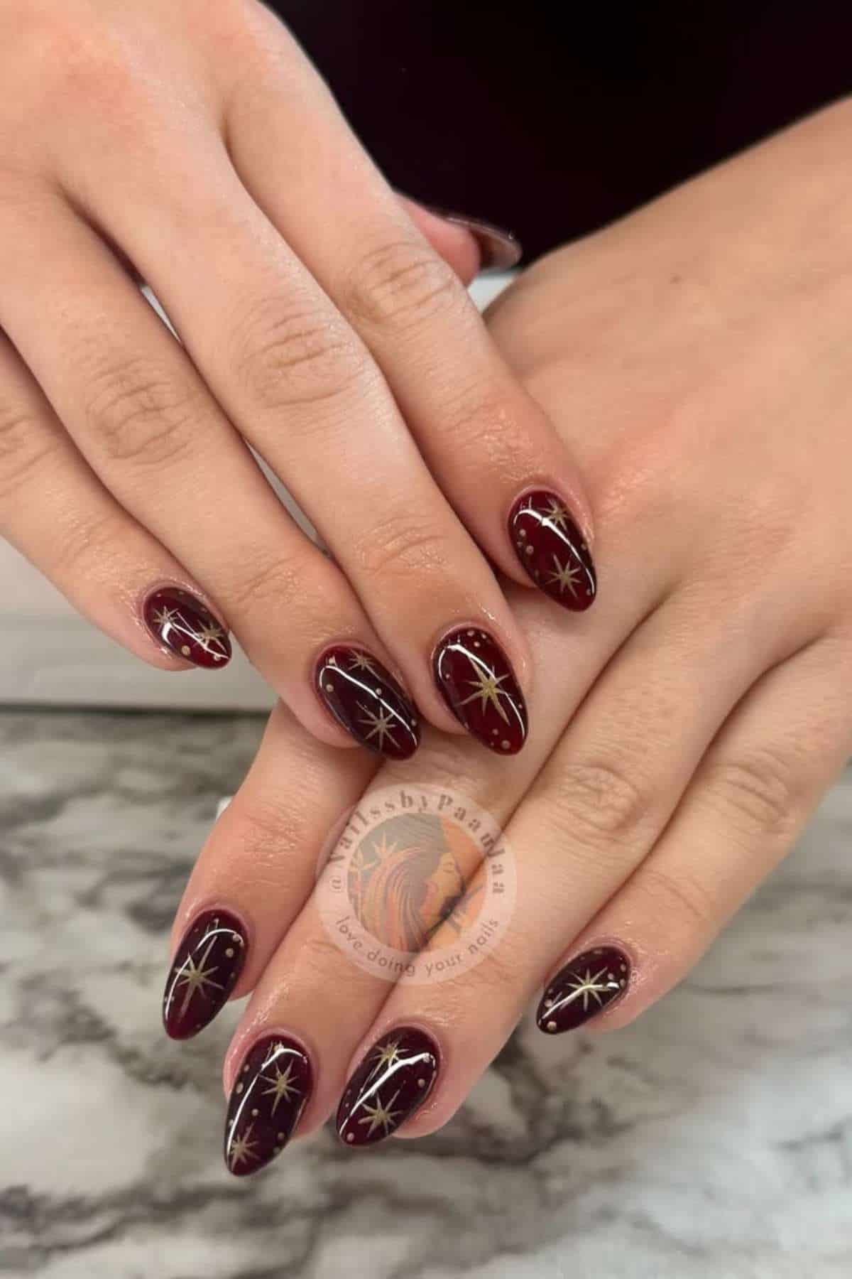 Hands with almond-shaped maroon nails featuring gold starburst designs, shown resting on a marble surface.