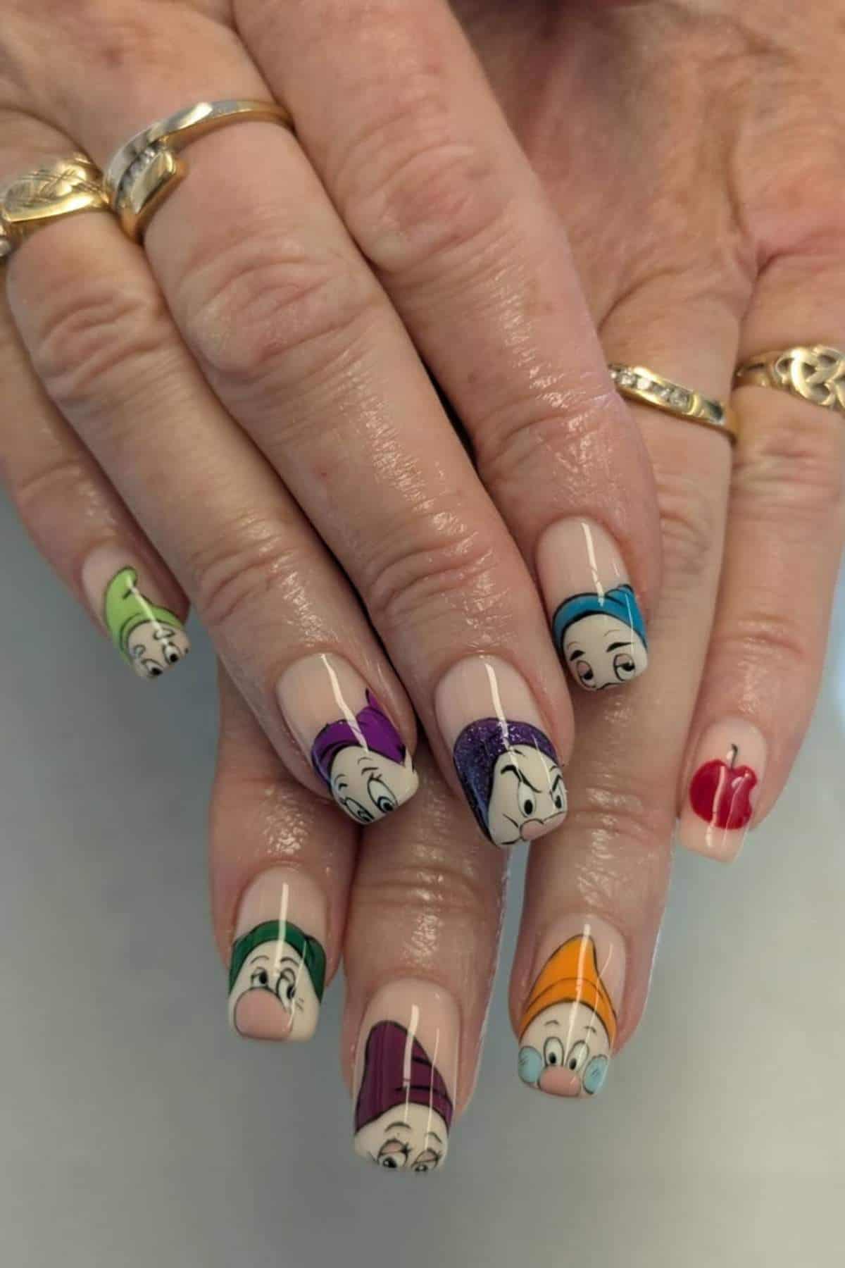 A hand with manicured nails featuring illustrations of the Seven Dwarfs and a red apple from Snow White, wearing gold rings.