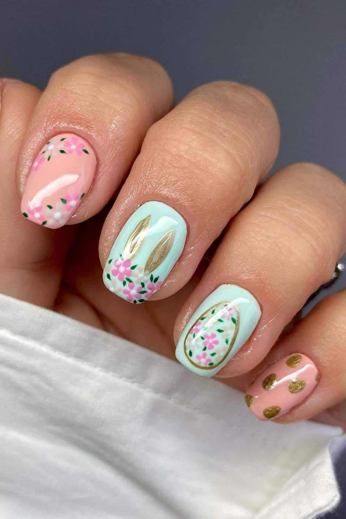 A hand with pastel pink and blue nail polish, decorated with floral patterns, gold bunny ears, and gold dots on the ring finger.