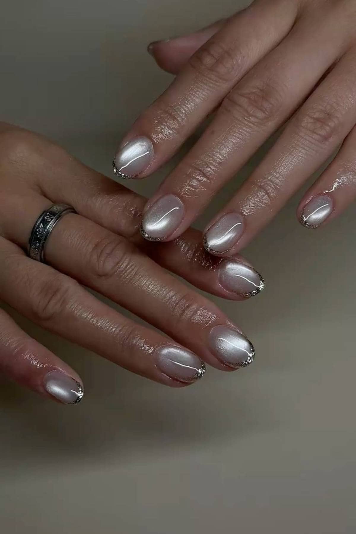 A pair of hands with short, oval nails painted in a pearly white polish and decorated with silver tips and small rhinestones. One hand wears a silver ring.