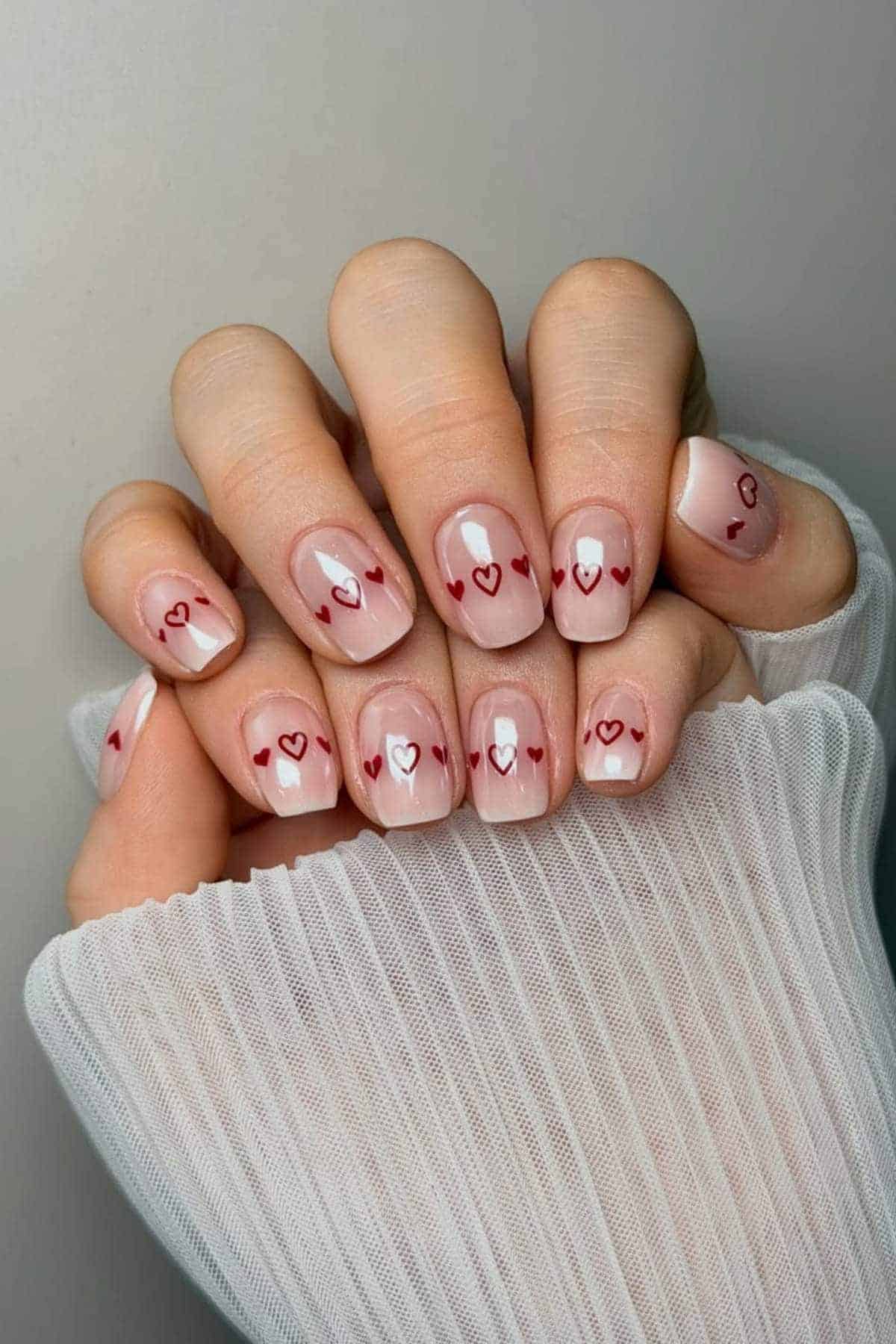 A hand with short, nude-colored nails featuring small red and white heart designs; the person is wearing a white, pleated, long-sleeve top.