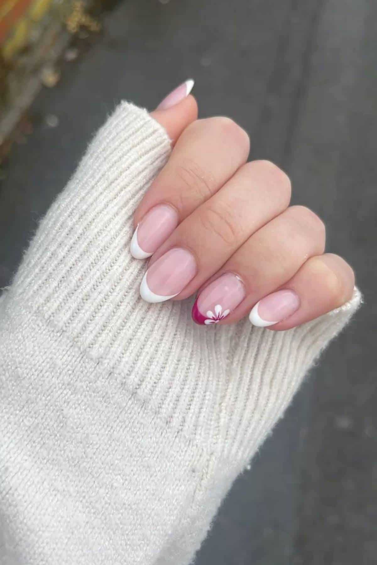 A hand with a white sweater sleeve shows nails painted with a French manicure, except one accent nail featuring a pink tip and a white flower design.