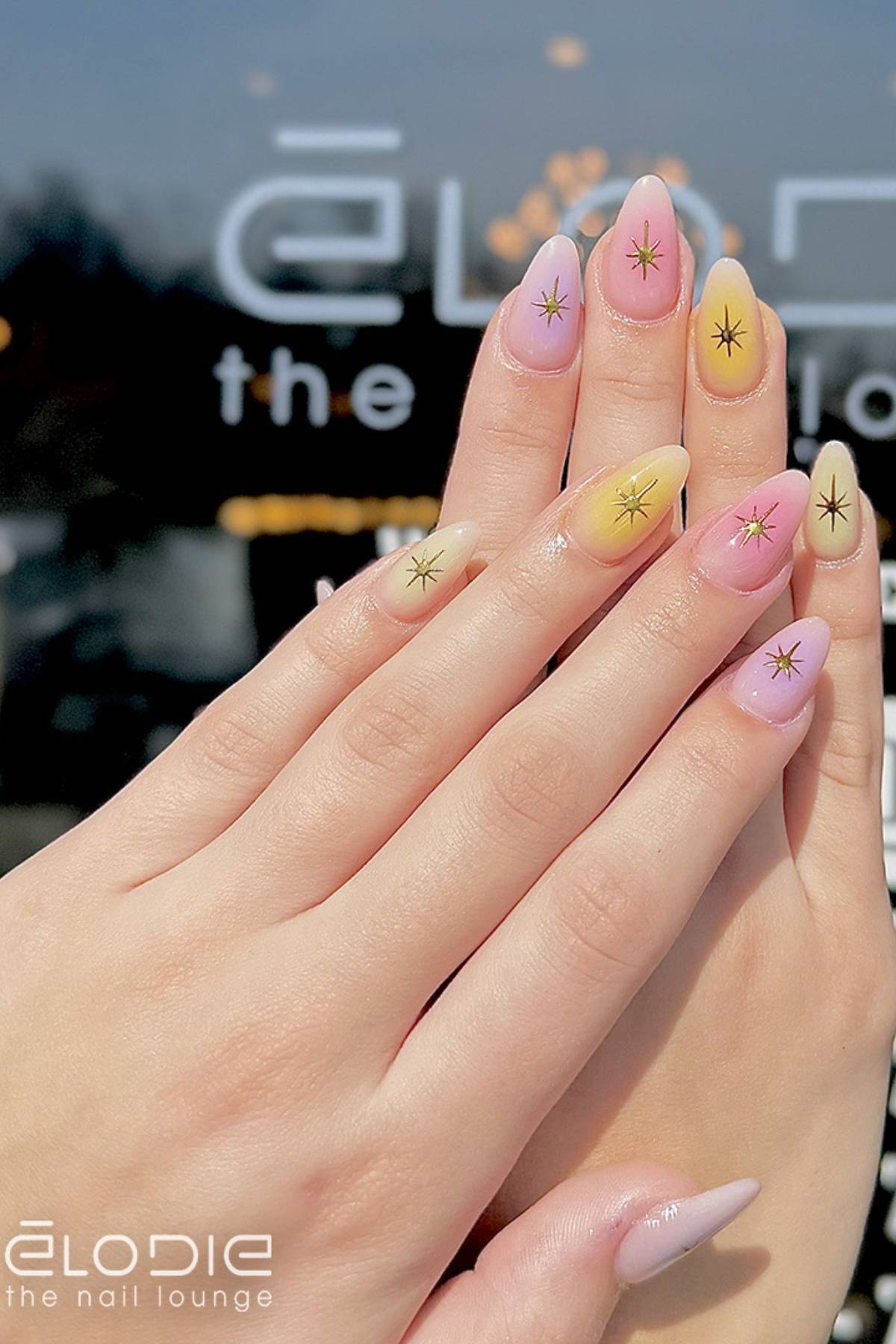 Close-up of hands with almond-shaped nails painted in pastel pink and yellow ombre, featuring small starburst designs; Elodie the Nail Lounge logo is visible in the background.