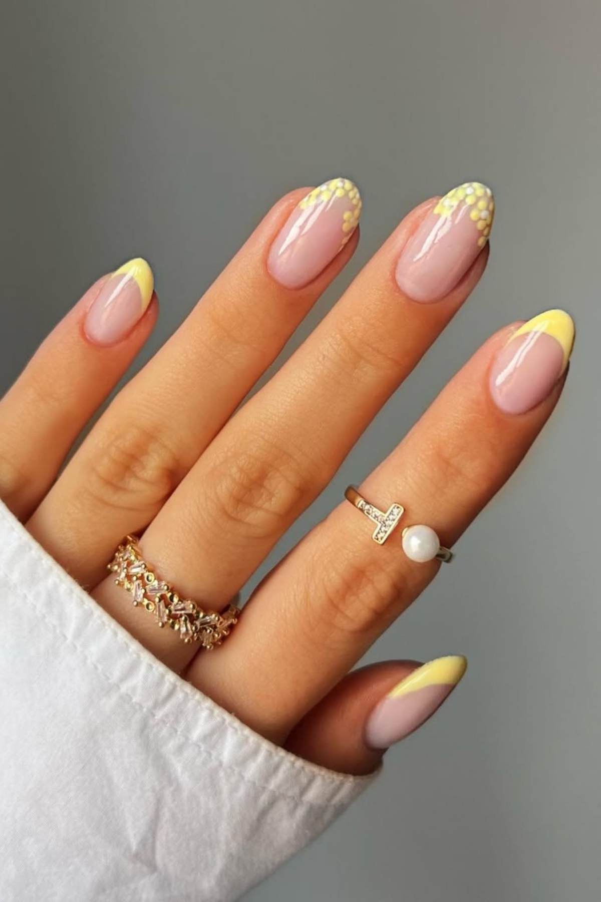 A hand with almond-shaped nails featuring a light pink base, yellow French tips, and small yellow dot accents. The person is wearing two rings and a white long-sleeve shirt.