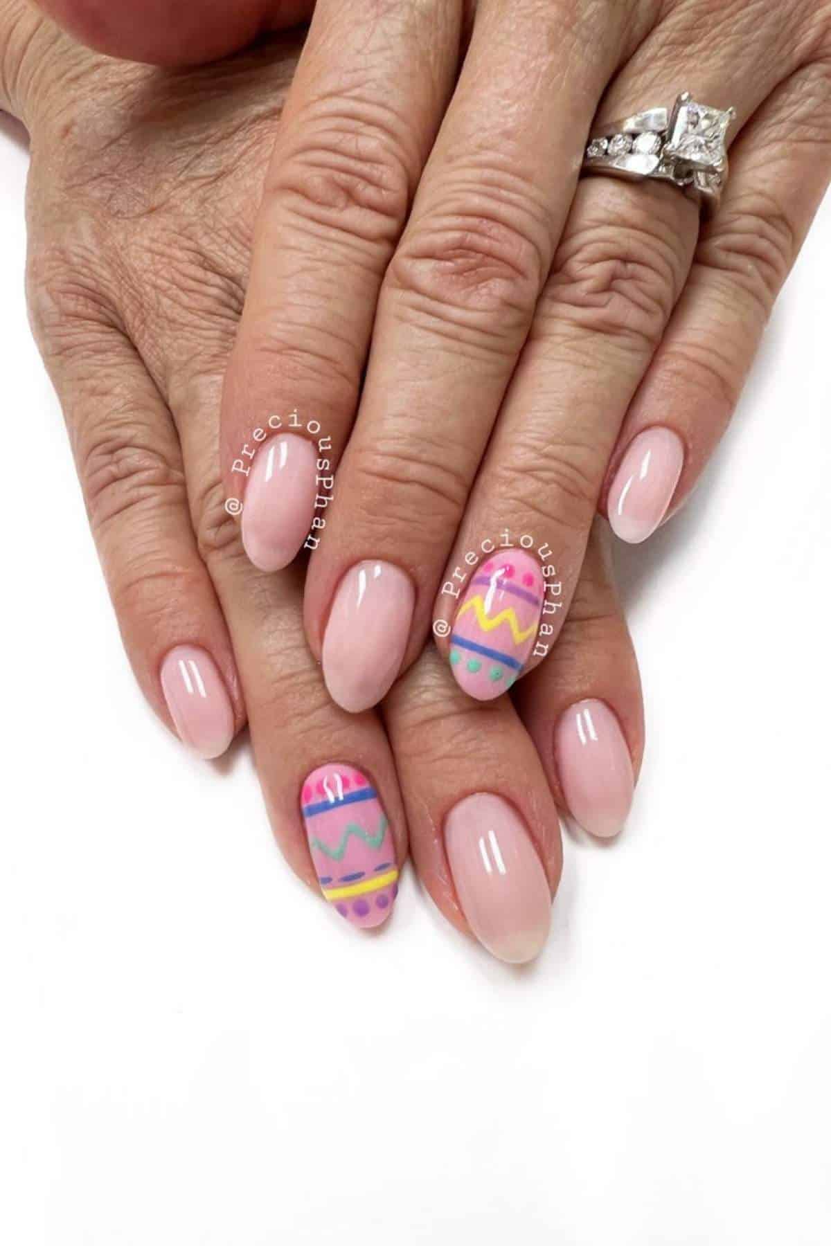 A hand with pale pink, almond-shaped nails; two nails feature colorful Easter egg designs in pink, yellow, blue, and green patterns.