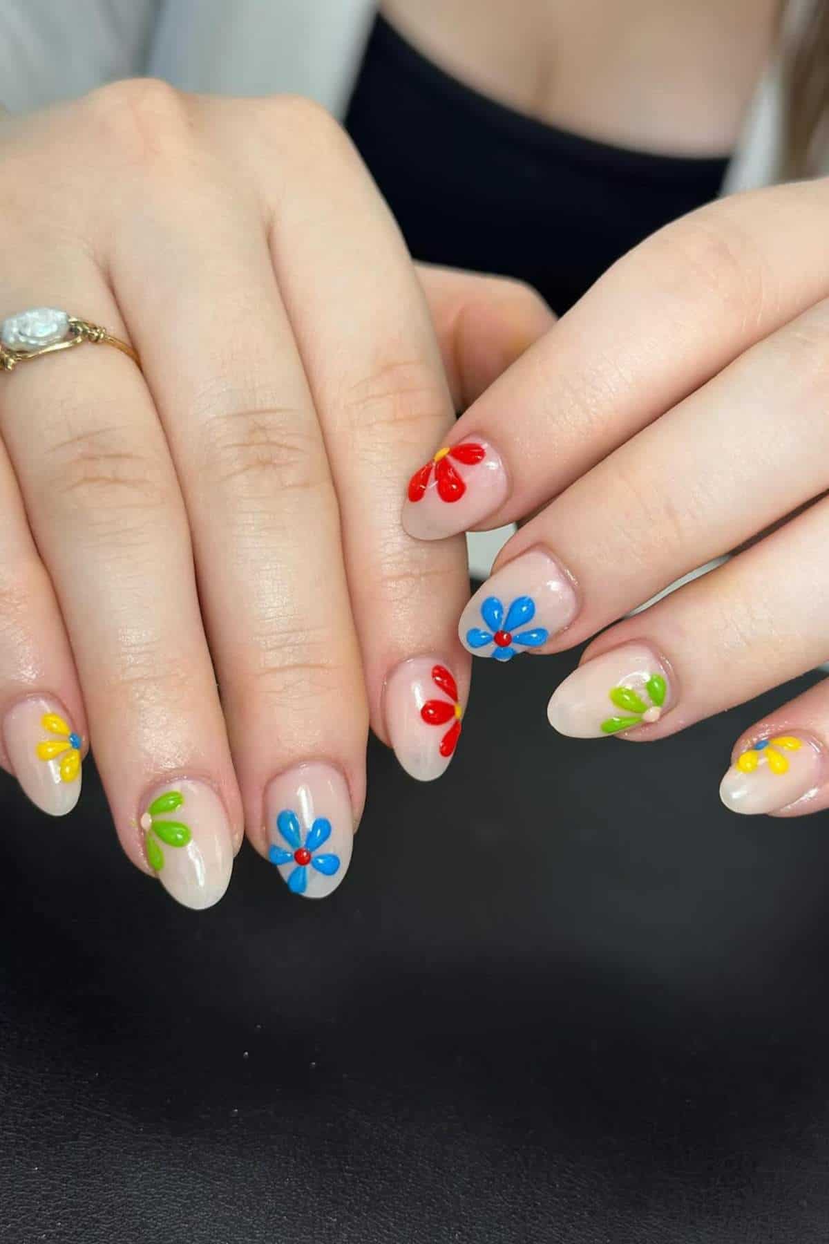 Hands with almond-shaped nails featuring colorful flower designs in red, blue, yellow, and green on a nude base. A gold ring with a clear stone is worn on one finger.