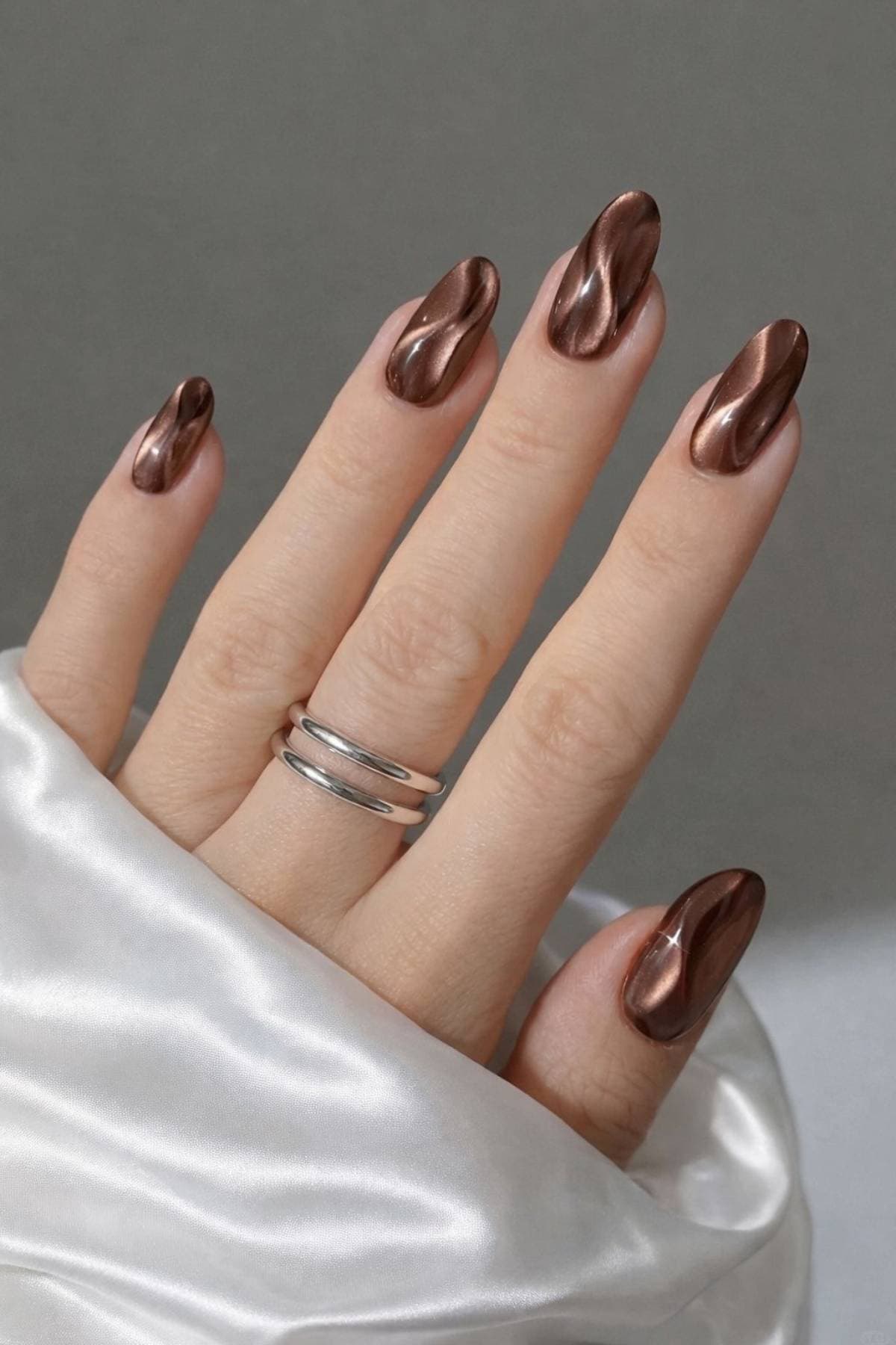 A hand with long, almond-shaped nails painted in metallic brown polish with a wavy, reflective design, wearing a simple silver ring, resting on white satin fabric.