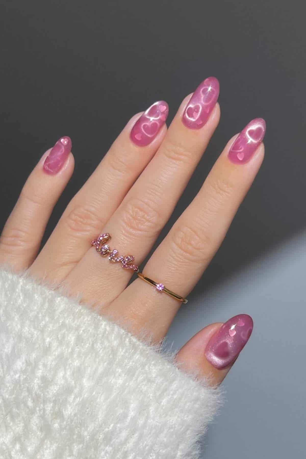 A hand with long, almond-shaped nails painted pink with shiny heart designs. The person is wearing two rings and a white fuzzy sweater.