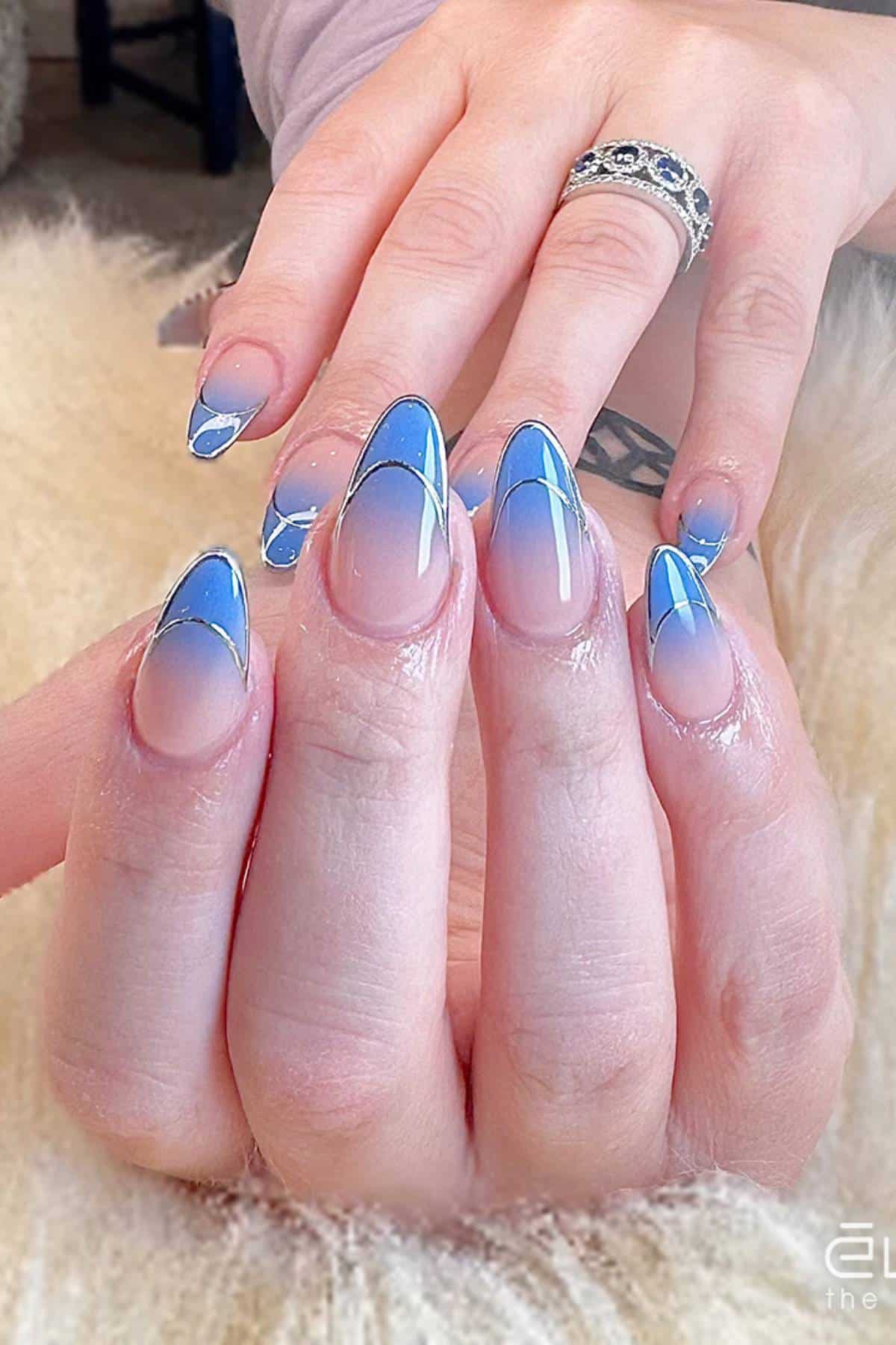 A hand with almond-shaped nails featuring a blue French tip design and white accent lines, with a silver ring on the index finger.
