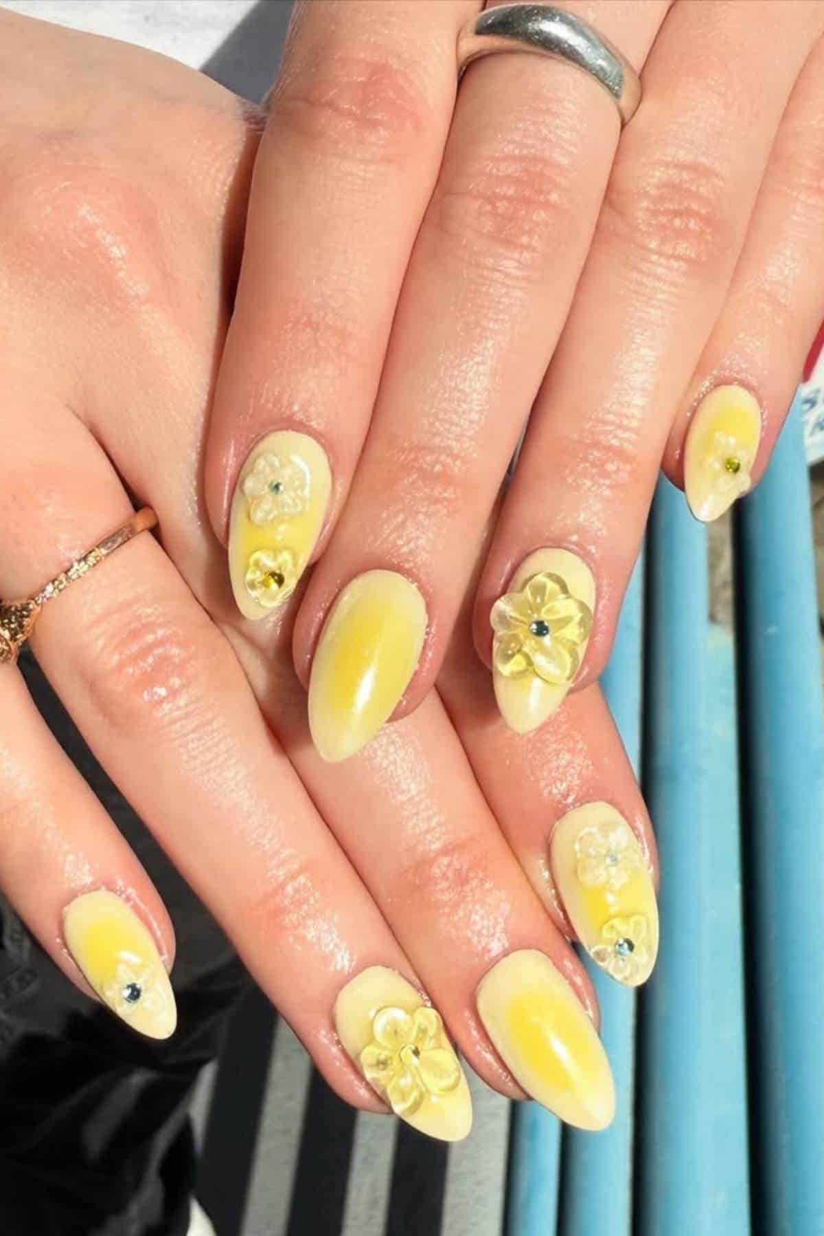 Hands with almond-shaped yellow ombre nails, decorated with 3D flower designs and rhinestones; person wears silver and gold rings.
