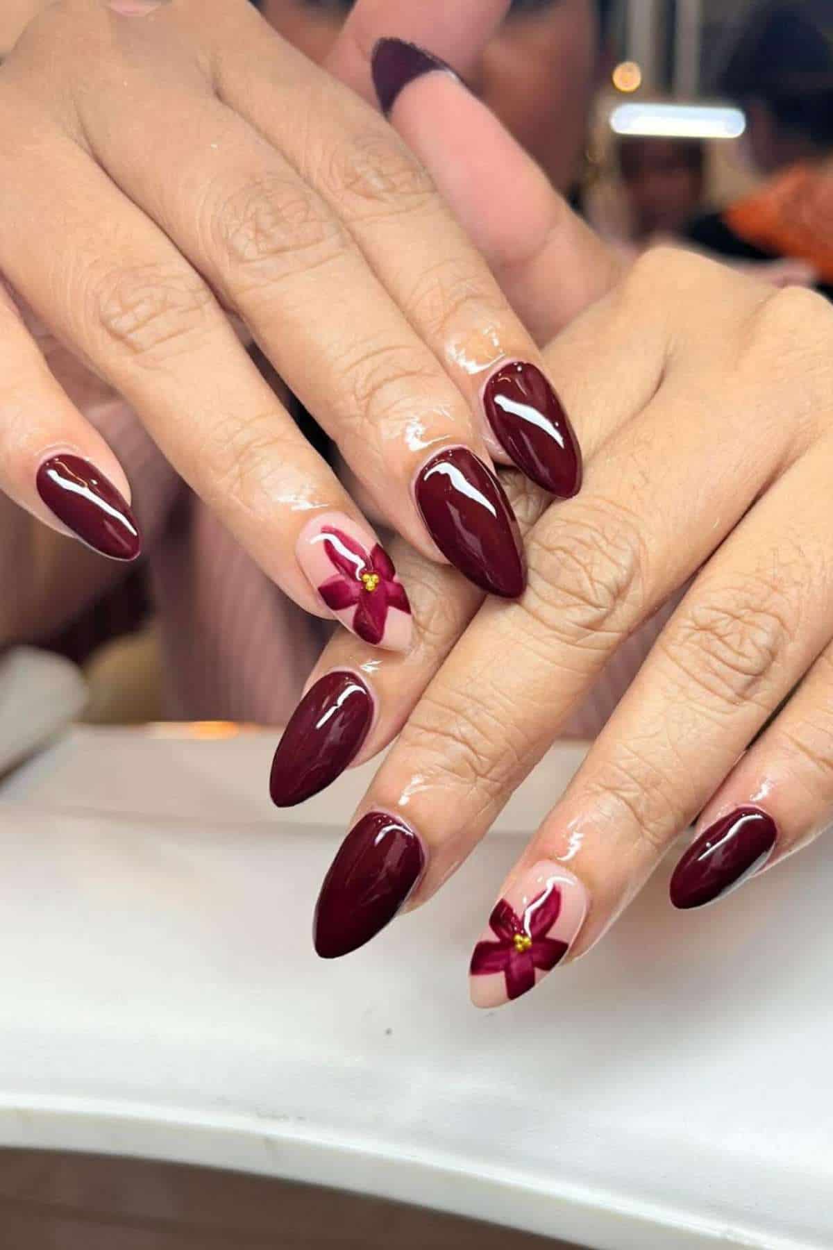 Hands with long, almond-shaped nails painted dark red; two nails feature a bow design with a small yellow accent.