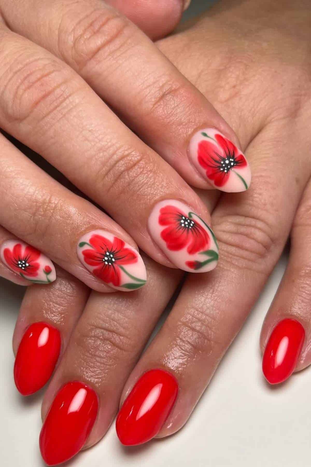 Hands with manicured nails; some nails are painted solid red while others feature red flower designs with green leaves and white accents.