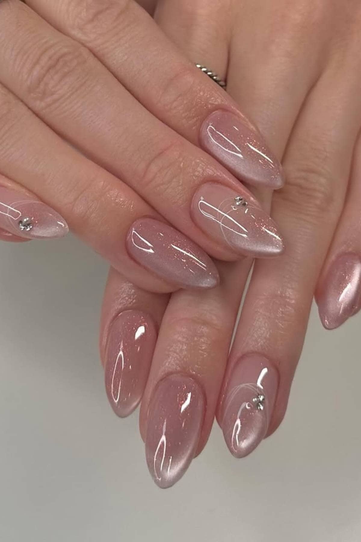 Hands with almond-shaped nails featuring a light pink ombre design, glossy finish, and small rhinestone accents on two nails.